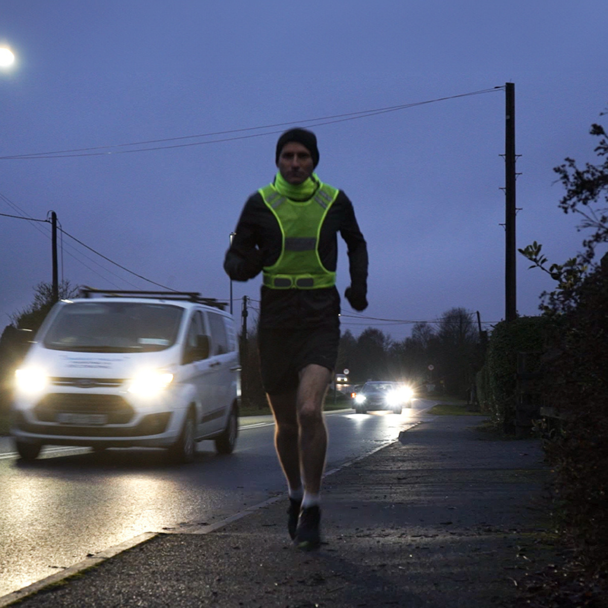 A runner jogging on a road at dusk wearing a Bodylite Gear Unisex V2 Reflective Vest Neon Yellow 01309-ALL SIZeS over a black top and shorts, with cars and streetlights visible in the background.