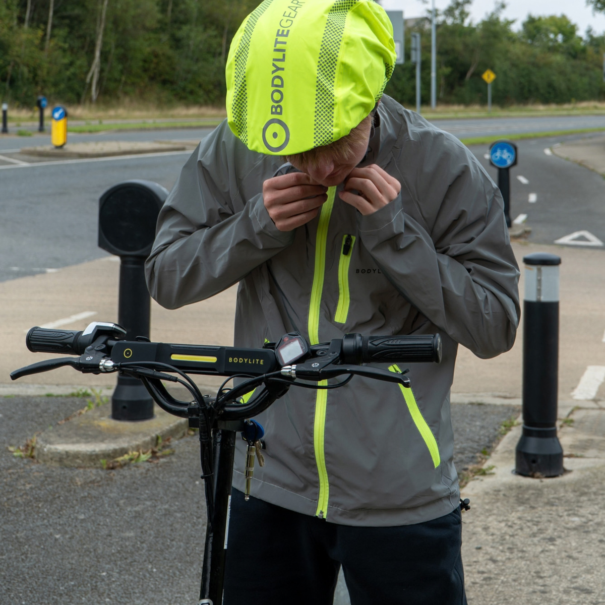 A person wearing a Bodylite Unisex Fully Reflective Jacket 01303-ALL SIZES and matching helmet stands rain cover beside a black electric scooter on a paved road, adjusting the helmet strap.