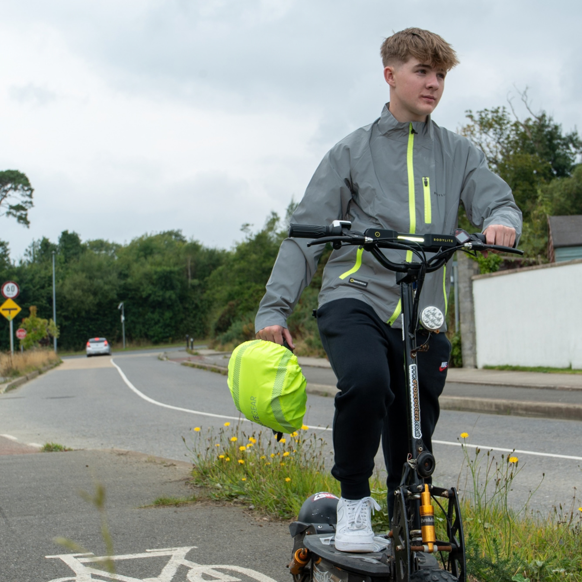 A young man riding an electric scooter on a road next to a bike lane, wearing a Bodylite Unisex Fully Reflective Jacket 01303-ALL SIZES, black trousers, and holding a bright yellow helmet rain cover.