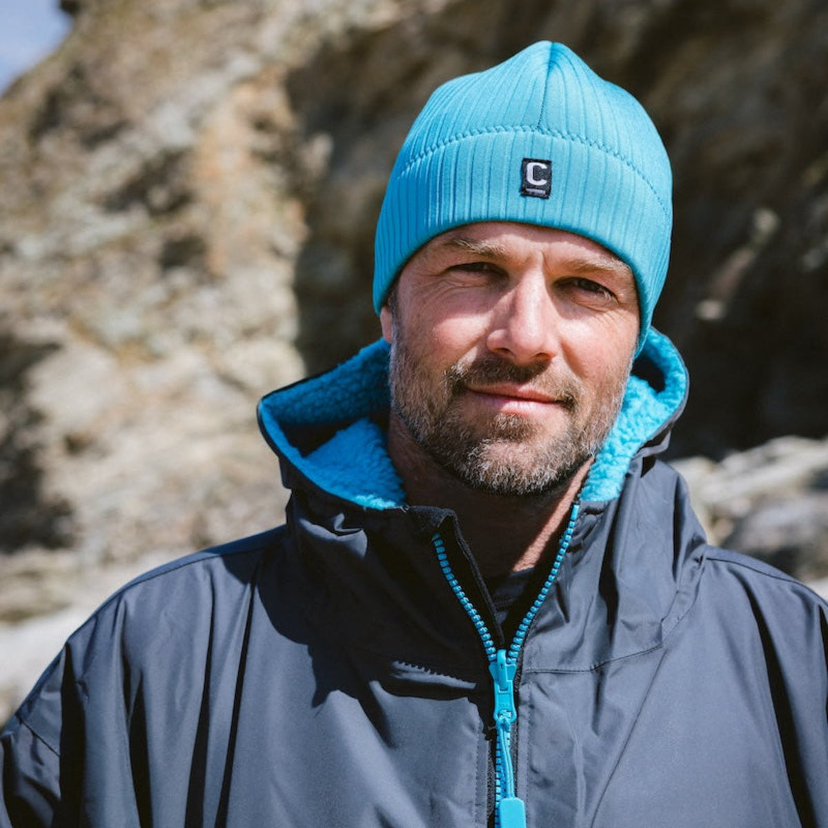 Man wearing a C-HOBE Unisex Storm Chaser Thermal Neoprene Swim Watersports Beanie Teal Blue and hardshell changing rob standing in front of a rocky background on a beach
