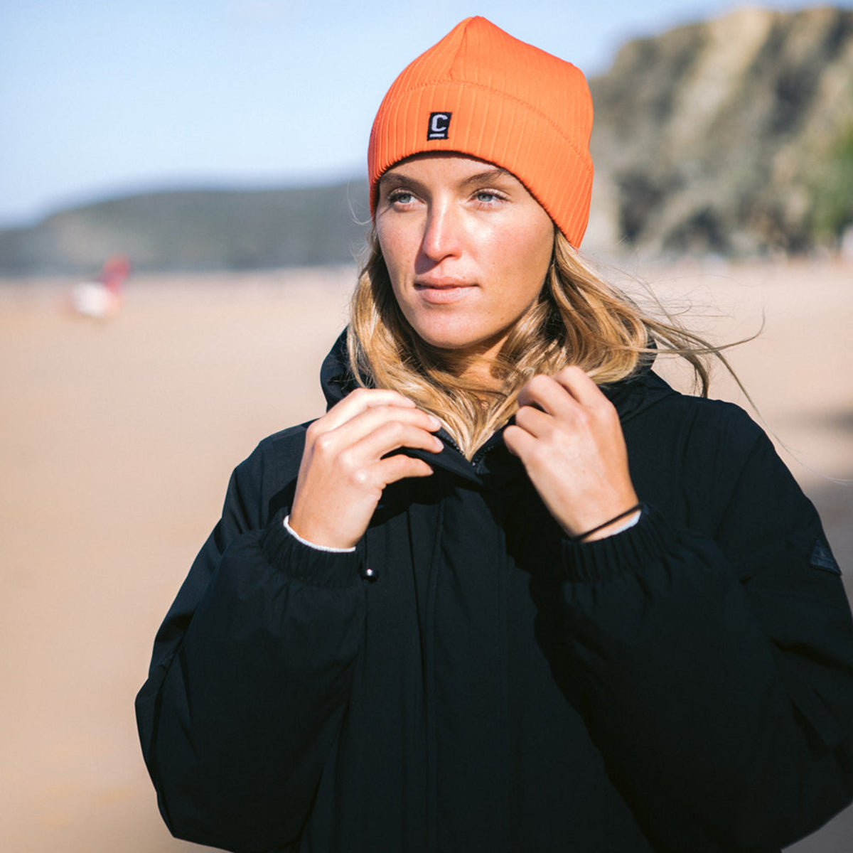 Woman wearing a C-Skins Unisex Storm Chaser Thermal Neoprene Swim Watersports Beanie Warm Red C-HOBE and a black changing robe on the beach