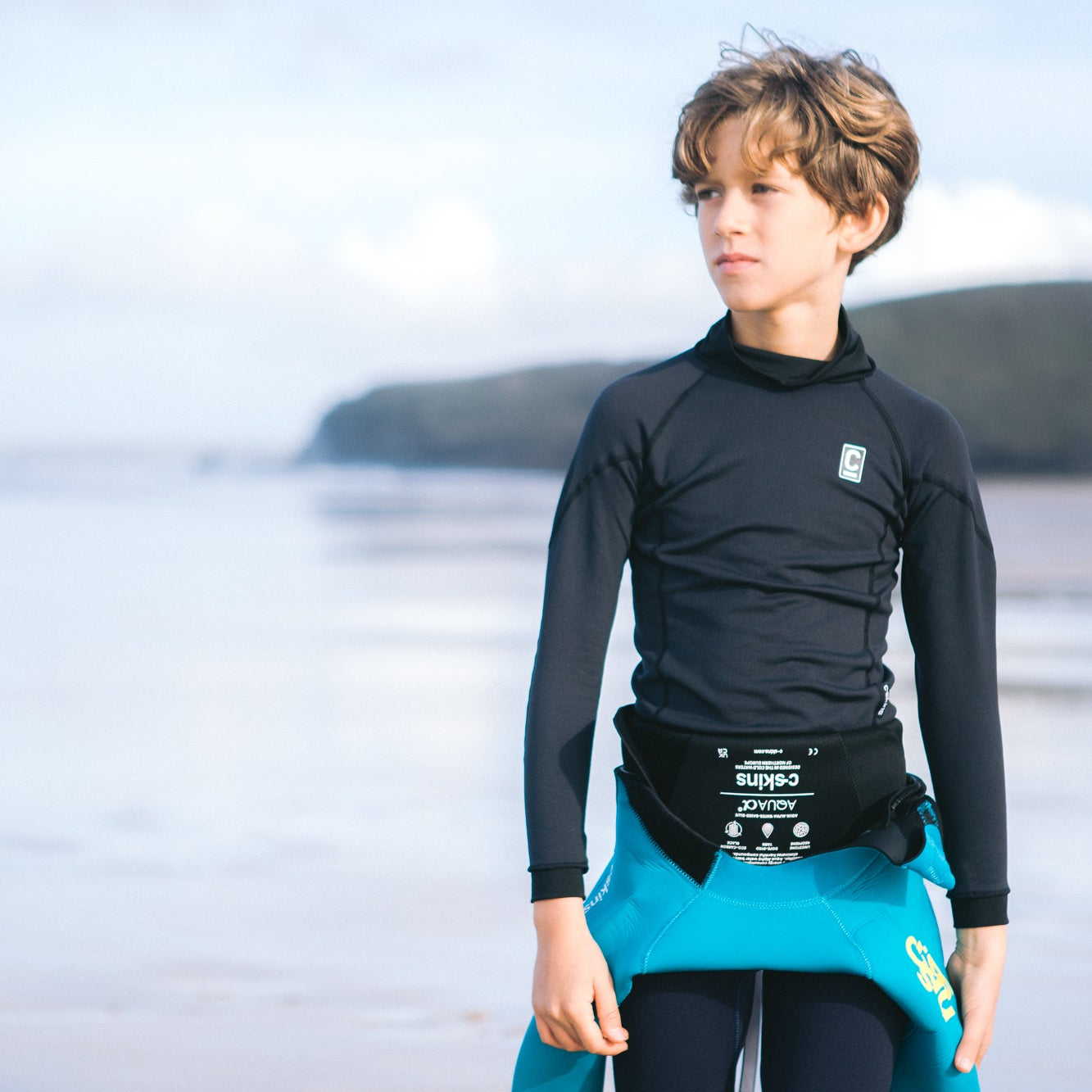 Kid wearing C-Skins Kids Junior Thermal Long Sleeve HDi Rash Vest Black C-HDILSJ with a blurred beach background