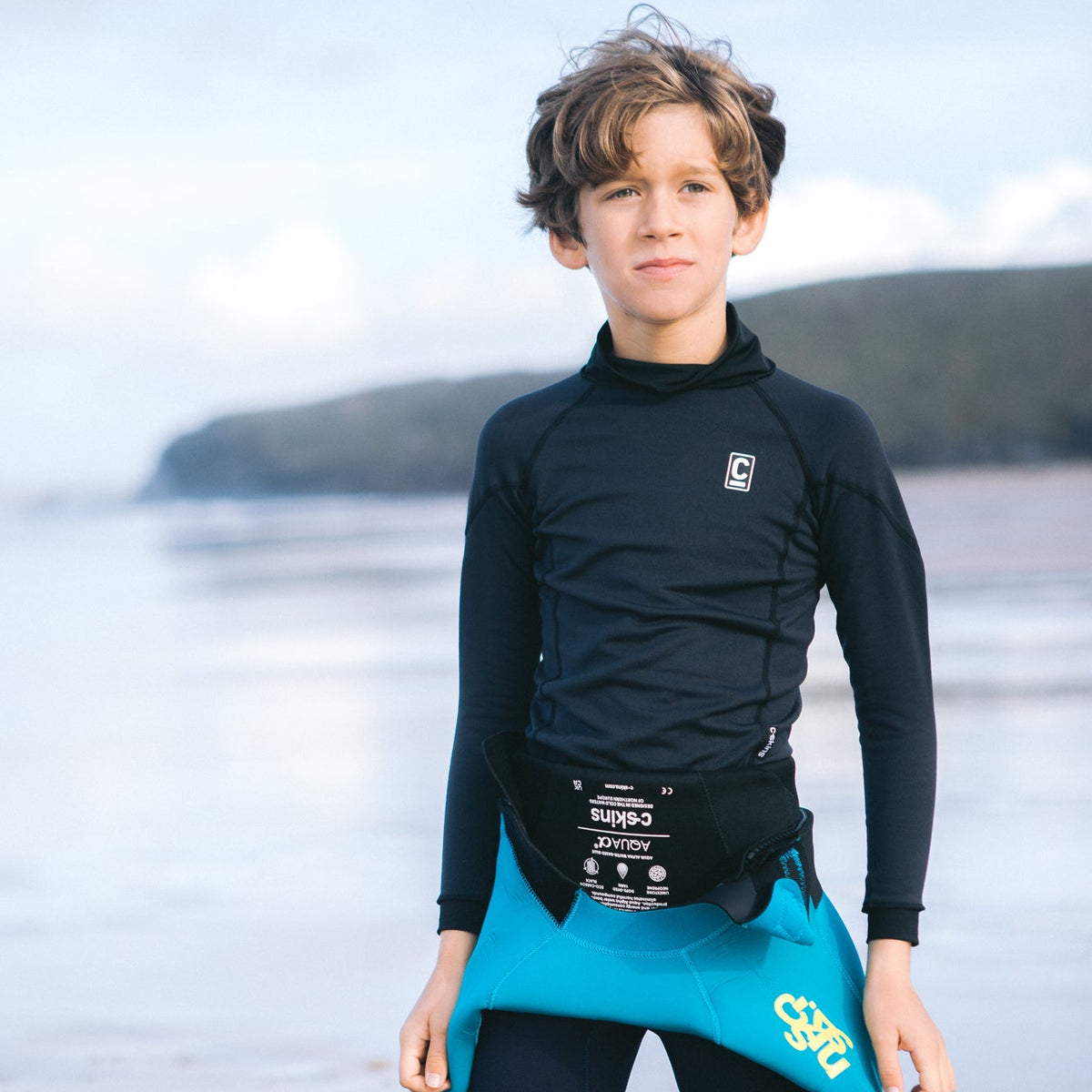 Kids wearing C-Skins Kids Junior Thermal Long Sleeve HDi Rash Vest Black C-HDILSJ with a blurred beach background