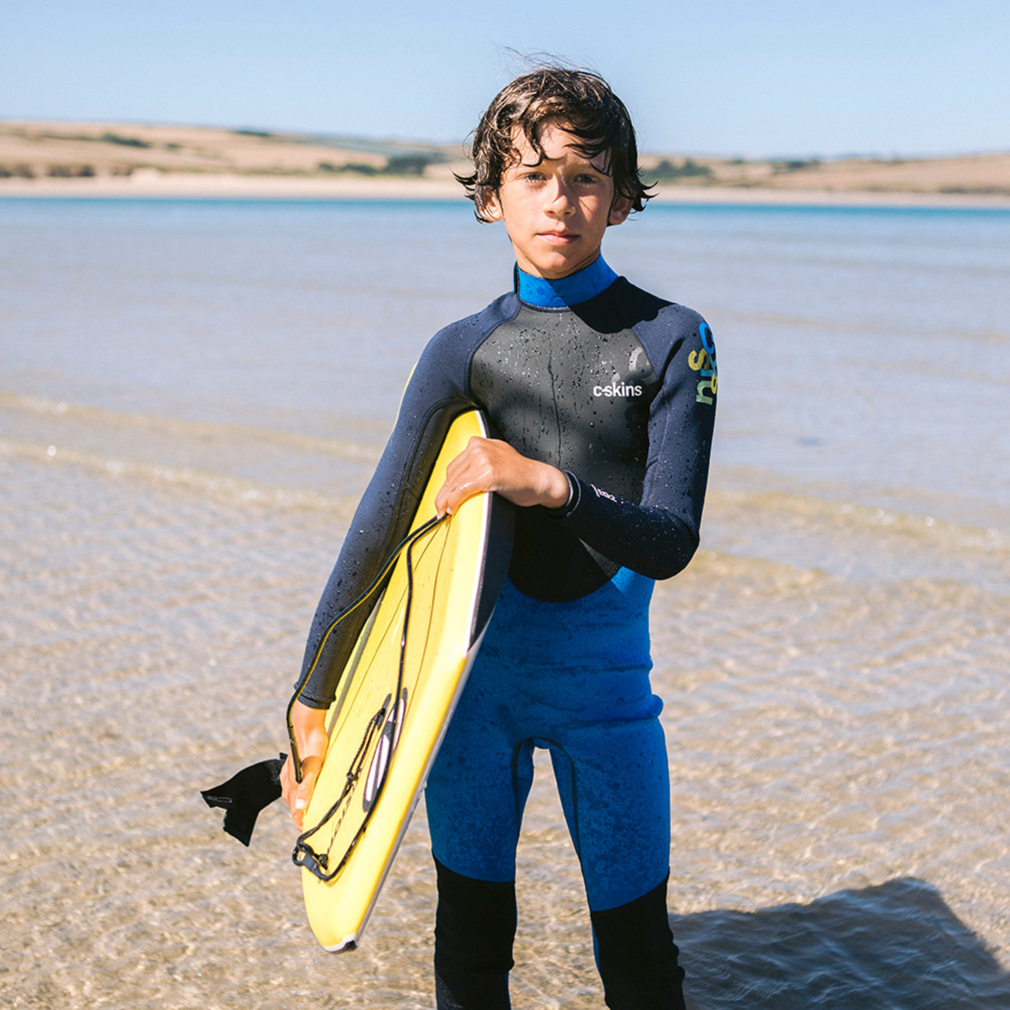 Kid wearing C-Skins Junior Unisex Element 3mm 2mm Flatlock Steamer Wetsuit Cyan Slate Multi C-EL32STJ holding a bodyboard on the beach