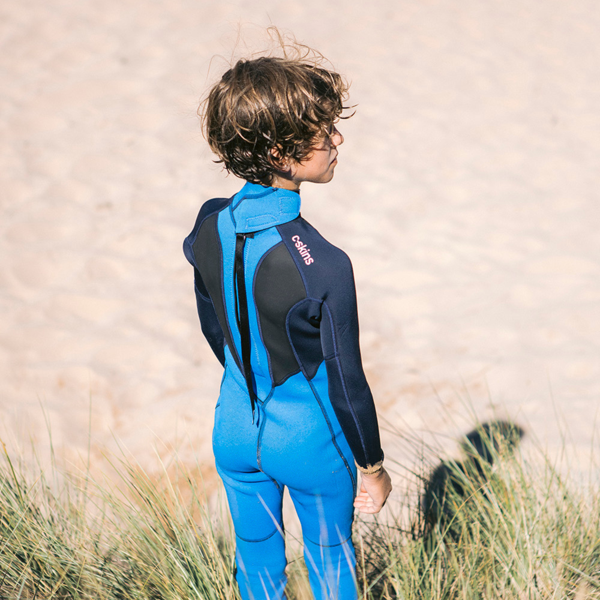 Kid wearing C-Skins Junior Unisex Element 3mm 2mm Flatlock Steamer Wetsuit Cyan Slate Multi C-EL32STJ on a beach with the back zip visible