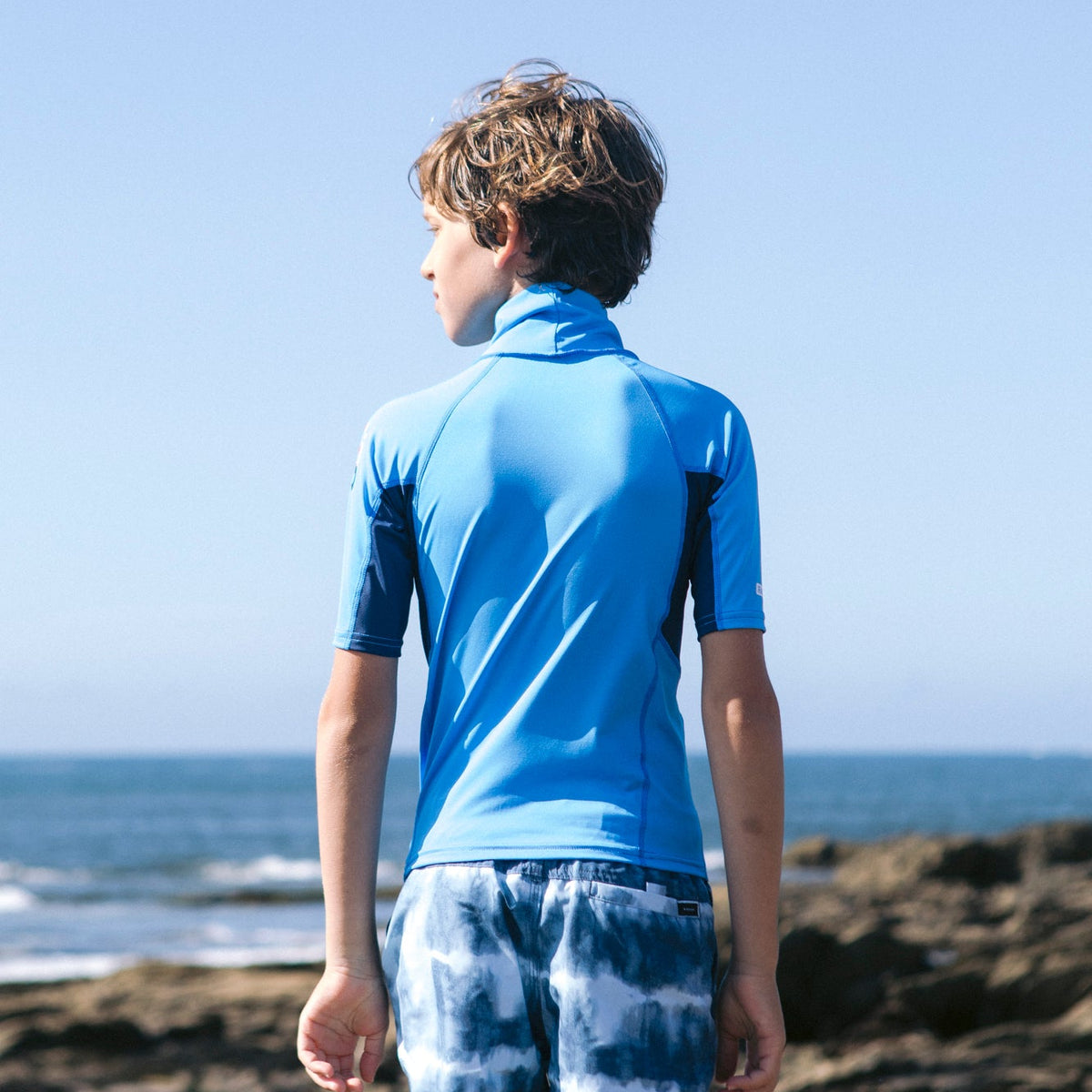 Boy wearing C-Skins Junior Unisex NuWave Rash X Short Sleeve UPF50 Rash Vest Cyan Slate Multi C-NLYSSJ, view of the back of the rash vest, with a blurred beach background