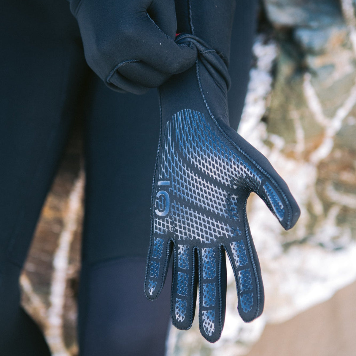 Person pulling on a C-Skins Unisex Legend Thermal Swim Watersports Neoprene Gloves 3mm C-GLLE, wearing a wetsuit, on the beach