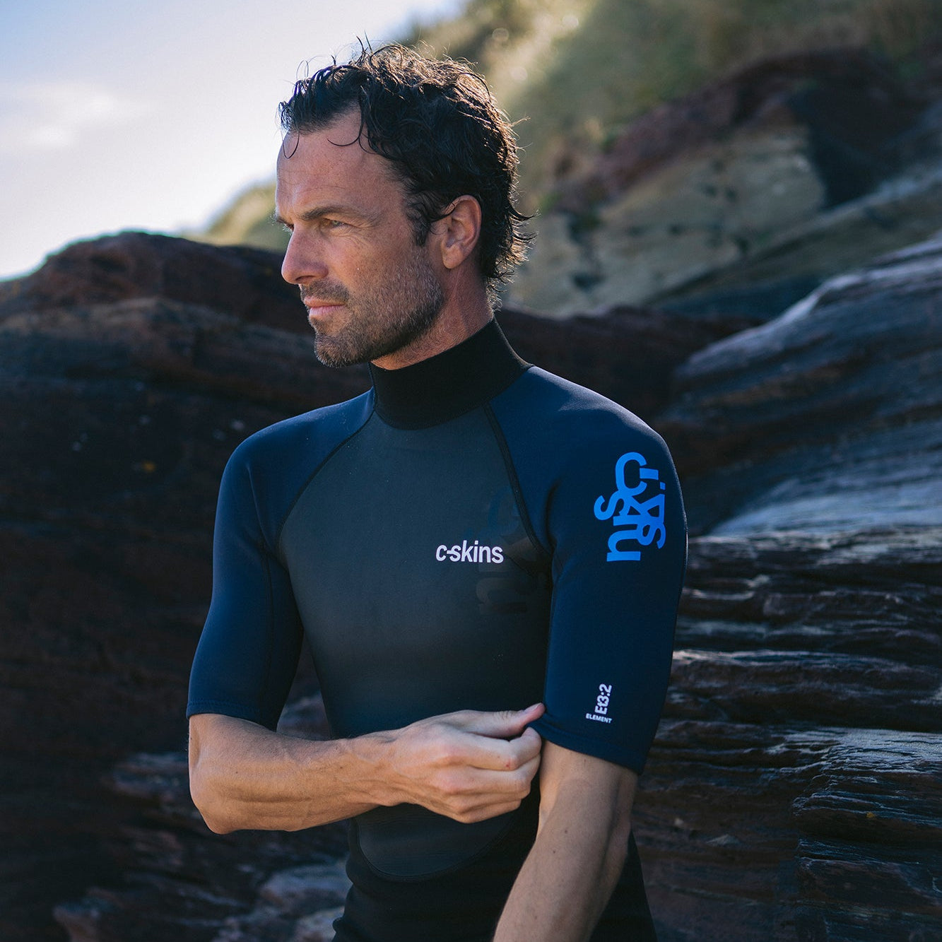 Close up of a man sitting on rock at the beach wearing C-Skins Men's Element 3mm 2mm Shorty Wetsuit Black Slate Cyan C-EL32SHM