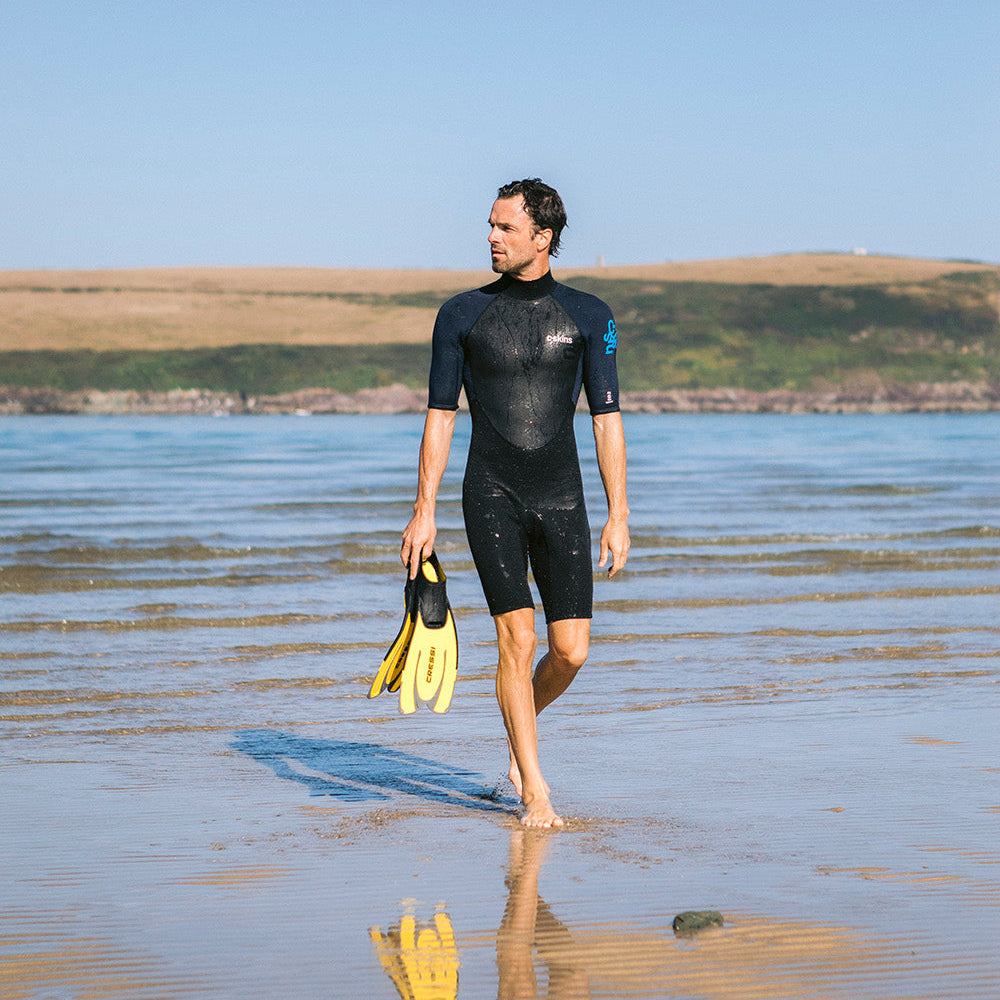 Man walking in the shallows on the beach wearing C-Skins Men's Element 3mm 2mm Shorty Wetsuit Black Slate Cyan C-EL32SHM holding flippers