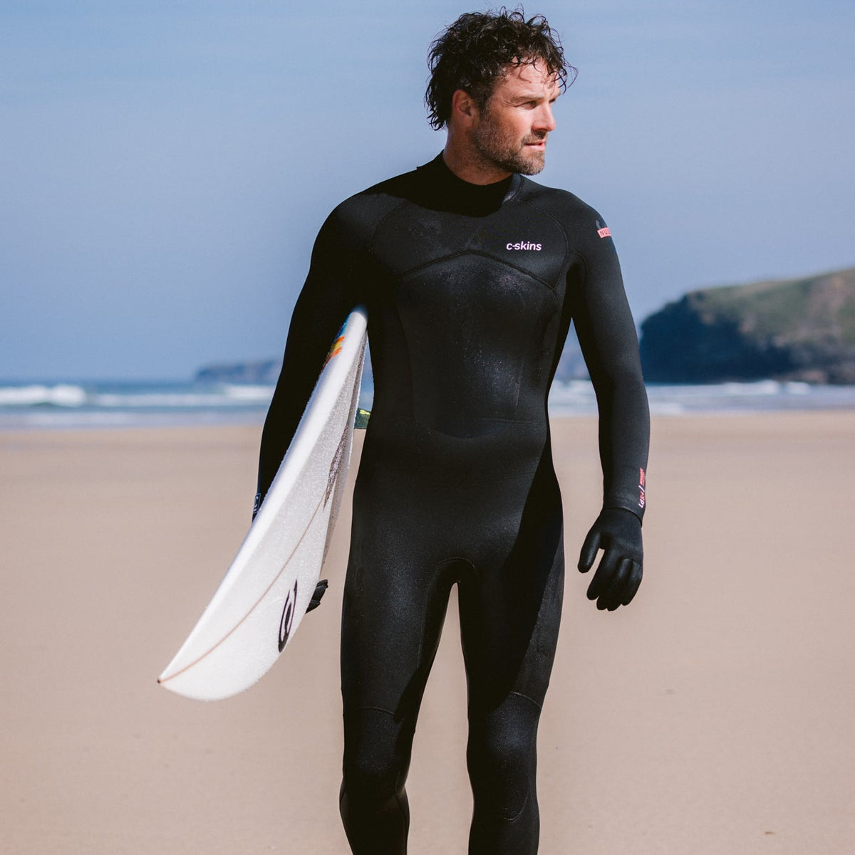 Man wearing C-Skins Men's NuWave Legend 5mm 4mm 3mm Back Zip GBS Steamer Black Neon Flare C-NLE54MBZ on the beach, carrying a surfboard
