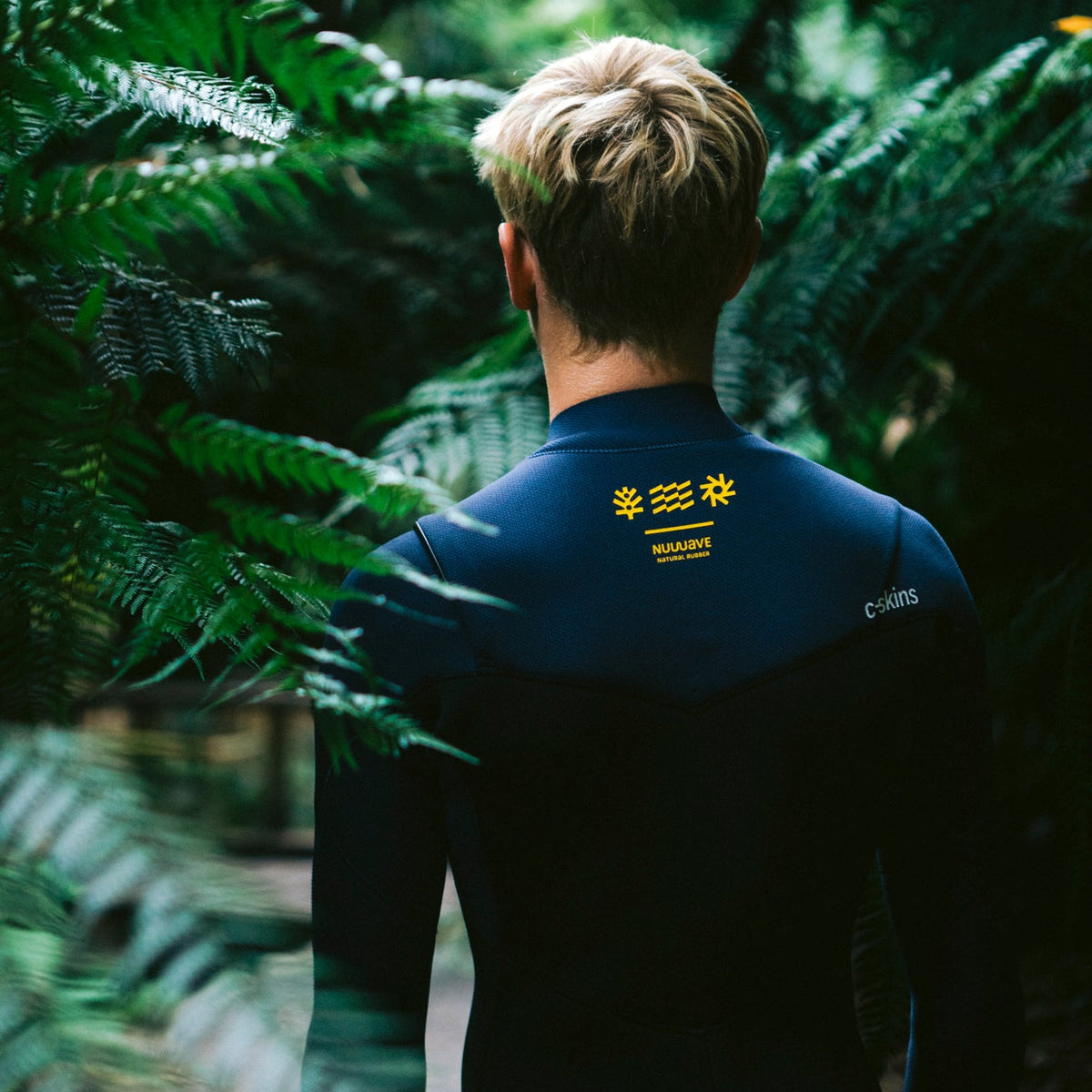 Man wearing a C-Skins Men's NuWave Session 4mm 3mm GBS Chest Zip Steamer Black Bluestone Saffron C-NSE43MCZ, shown front behind, standing amongst ferns