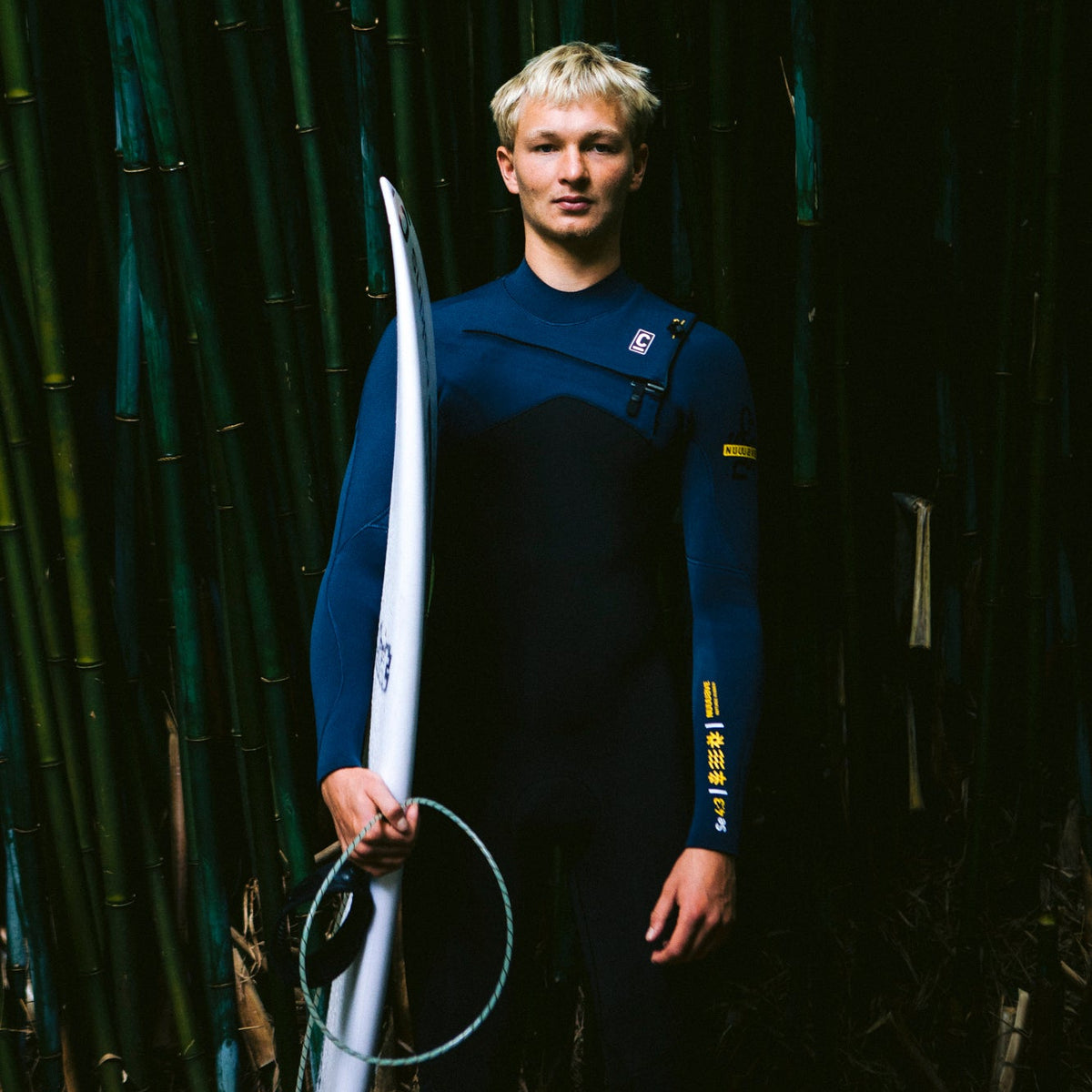 Man standing in front of bamboo, holding a surfboard, wearing a C-Skins Men's NuWave Session 4mm 3mm GBS Chest Zip Steamer Black Bluestone Saffron C-NSE43MCZ