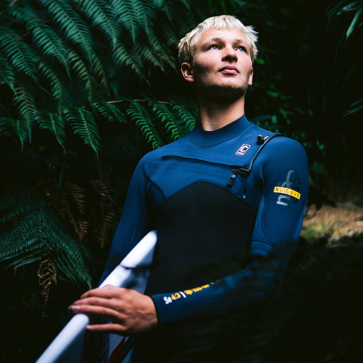Man wearing C-Skins Men's NuWave Session 4mm 3mm GBS Chest Zip Steamer Black Bluestone Saffron C-NSE43MCZ, holding a surfboard with ferns in the background