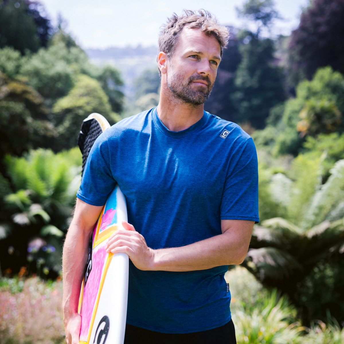 Mean wearing C-Skins Men's NuWave UV UPF50 Short Sleeve Surf Tee Deep Blue C-NLYSURFM, holding a surfboard, with a green grassy background