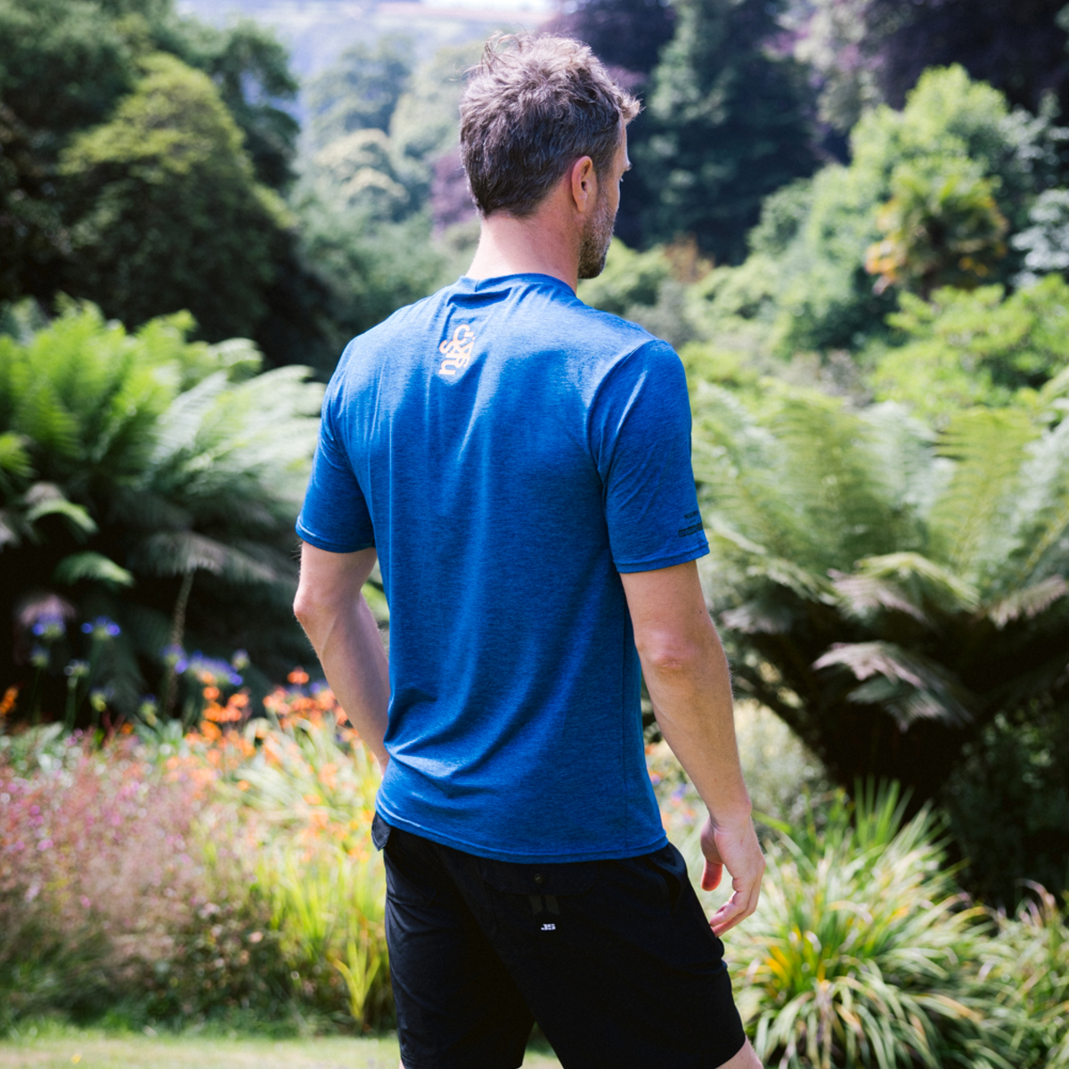 Man wearing C-Skins Men's NuWave UV UPF50 Short Sleeve Surf Tee Deep Blue C-NLYSURFM with a green grassy background. Back of rash vest is shown