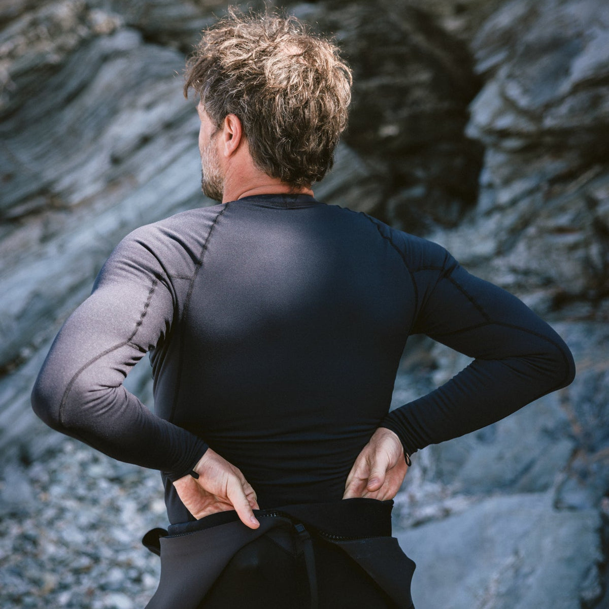 Person wearing a black wetsuit and C-Skins Men's HDi Thermal Long Sleeve Rash Vest Black C-HDILSMC standing on rocky terrain with his back turned