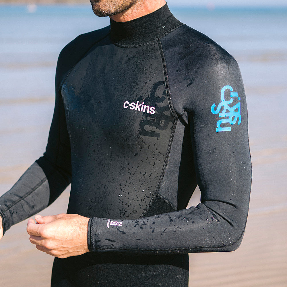 Close up of man wearing C-Skins Men's Element 3mm 2mm Flatlock Steamer Black Anthracite Cyan C-EL32STM with a blurry beach bakground