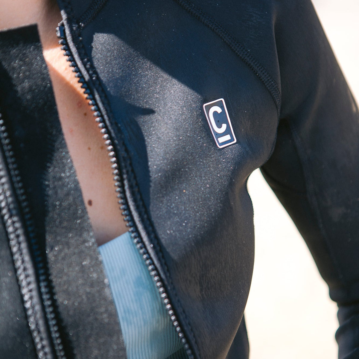 Close-up of a C-Skins Women's NuWave Solace Lite Flatlock Front Zip Long Sleeve Neoprene Jacket 1.5mm Black C-NSOL15VELS showing logo on a blurred background