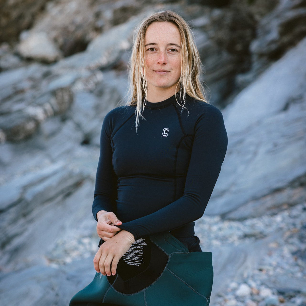 Woman wearing a black wetsuit and C-Skins Women's HDi Thermal Long Sleeve Rash Vest Black C-HDILSWC beside a rocky beach background