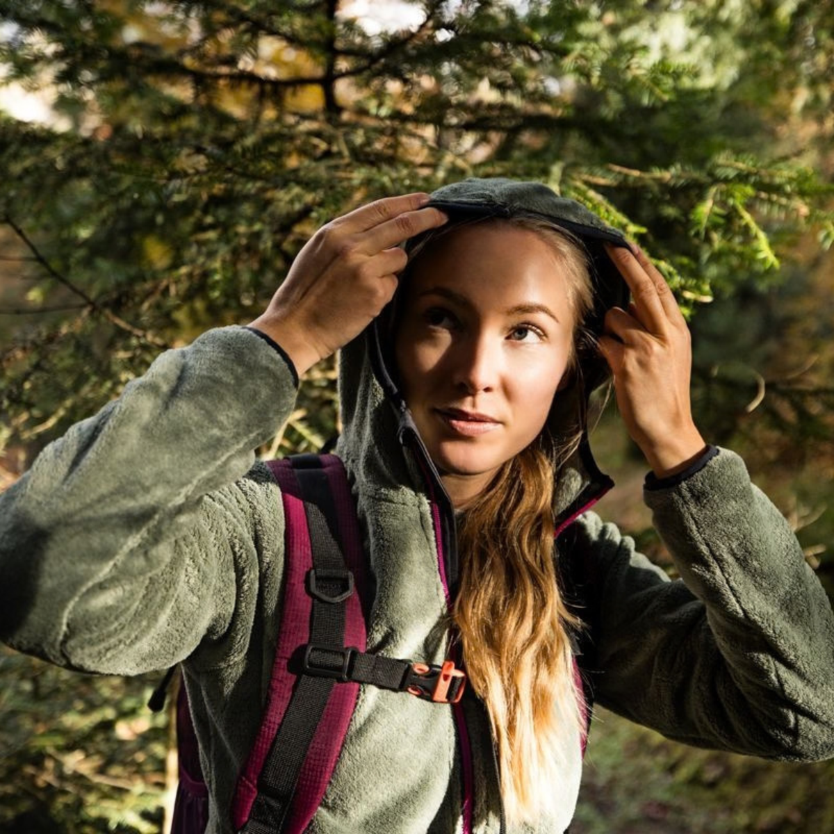 A woman outdoors in a forest setting, wearing a dark green fleece hooded jacket and adjusting the hood, with a CMP Laredo Trekking Daypack 22L C910 Amaranto 3V17957.