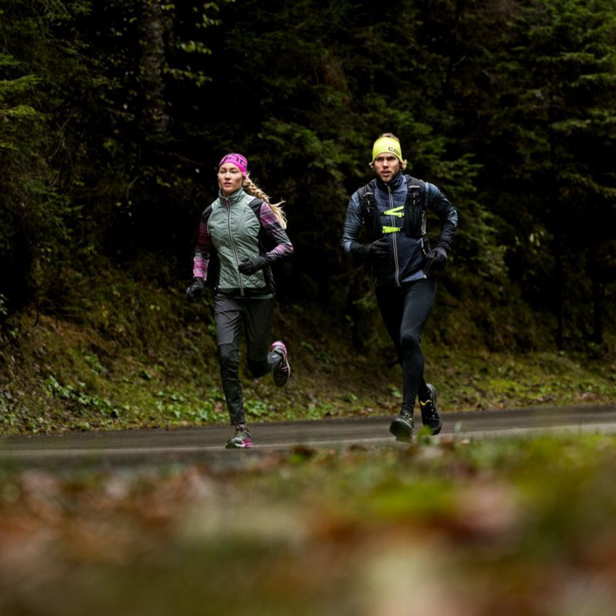 Two trail runners jogging on a forest road, with one wearing a sage green vest and pink headband and the other in a CMP Unisex Santa Fe Trail Running Vest Titanio U911 3V31537 and yellow headband.