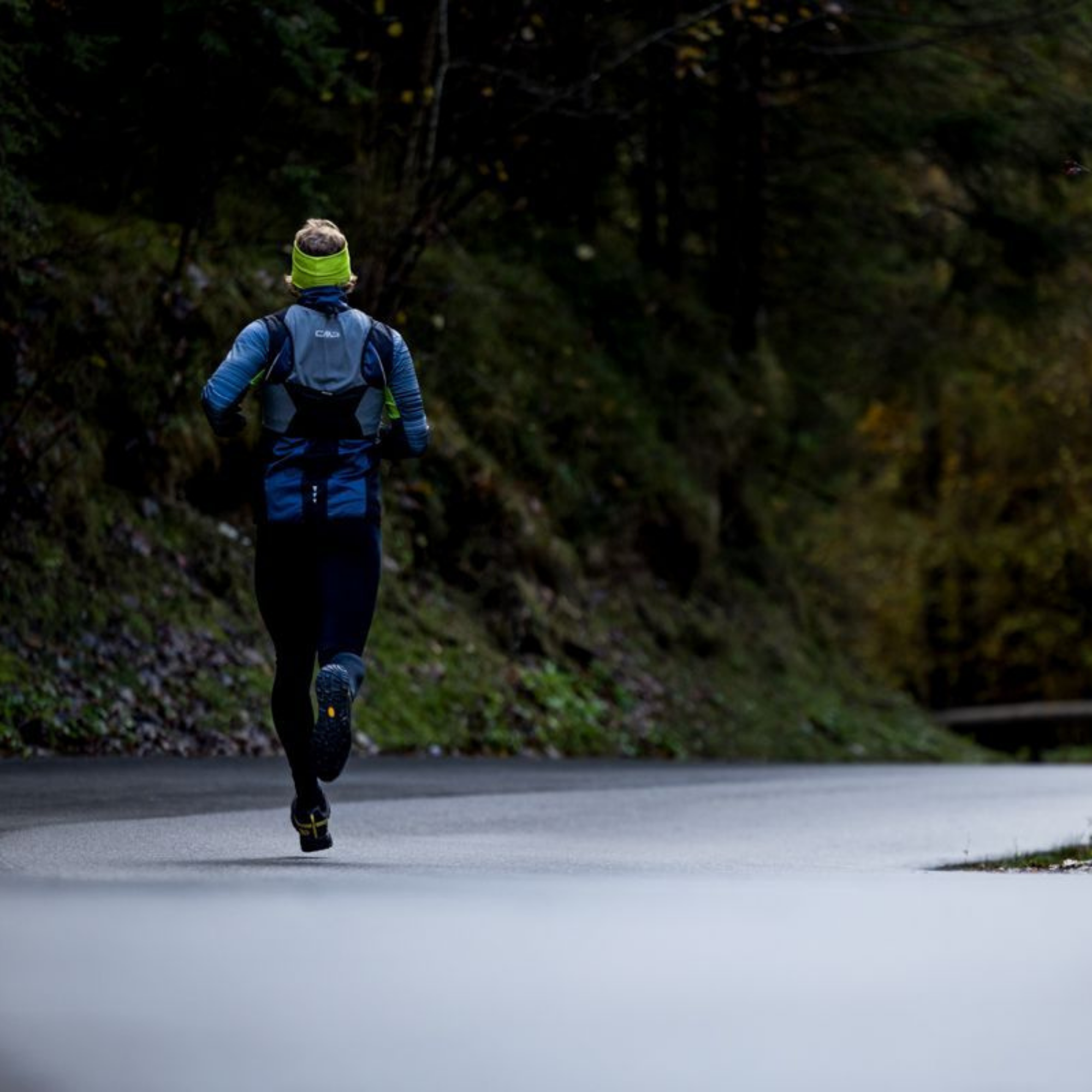 A trail runner viewed from behind running on a paved road through a forested outdoor setting. The runner wears a CMP Unisex Santa Fe Trail Running Vest Titanio U911 3V31537, a bright yellow-green headband, and dark running shoes.