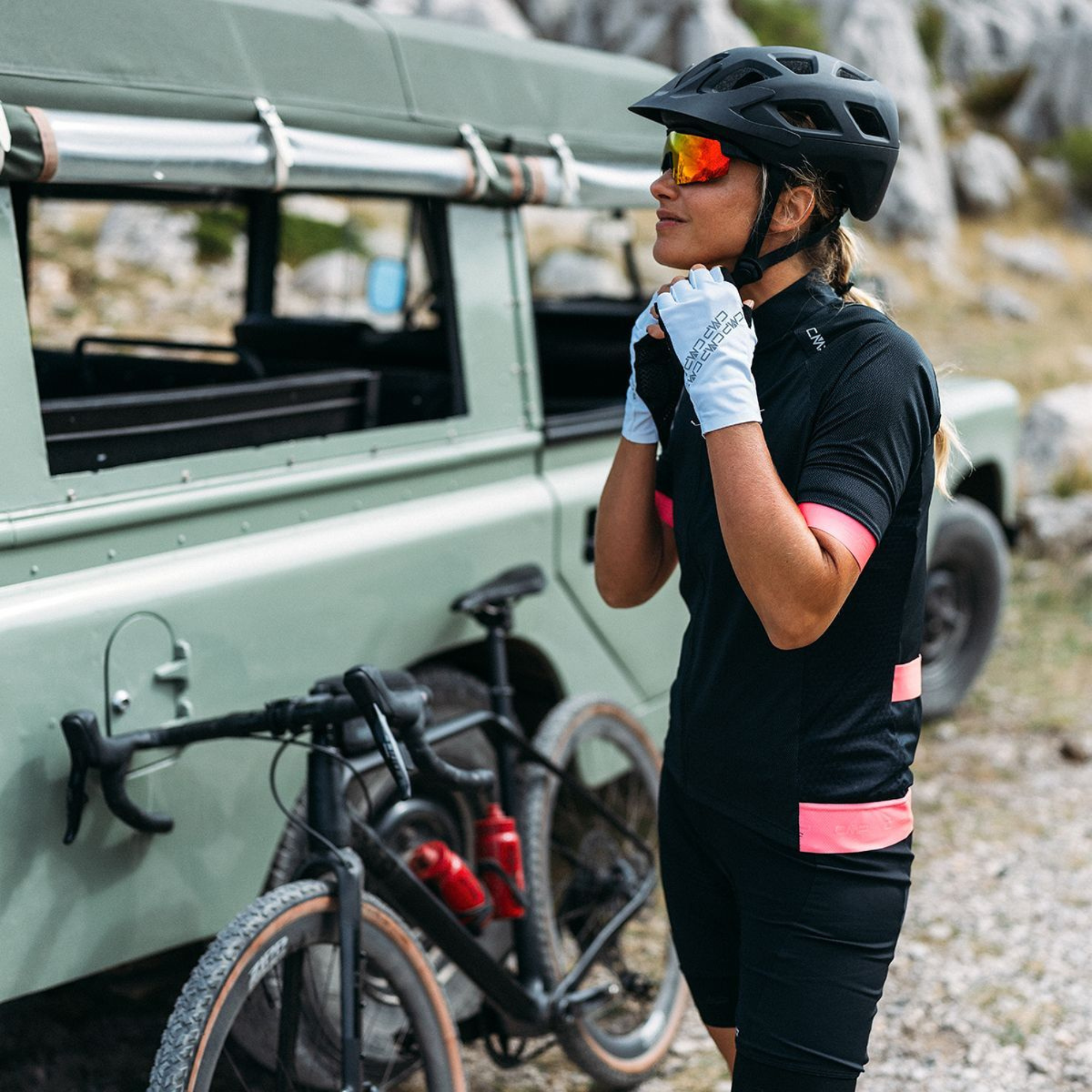 A woman wearing a black cycling jersey with pink accents, CMP Women's Bike Shorts with Sponge Pad Nero U901 34C7946, a black helmet, and white gloves stands next to a black bicycle parked in front of an olive green off-road vehicle outdoors.