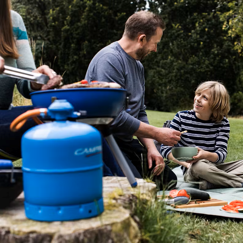 Camping Gaz 907 Gas Refill Only GAS907 connected to a camping stove while a family eats a picnic in the background