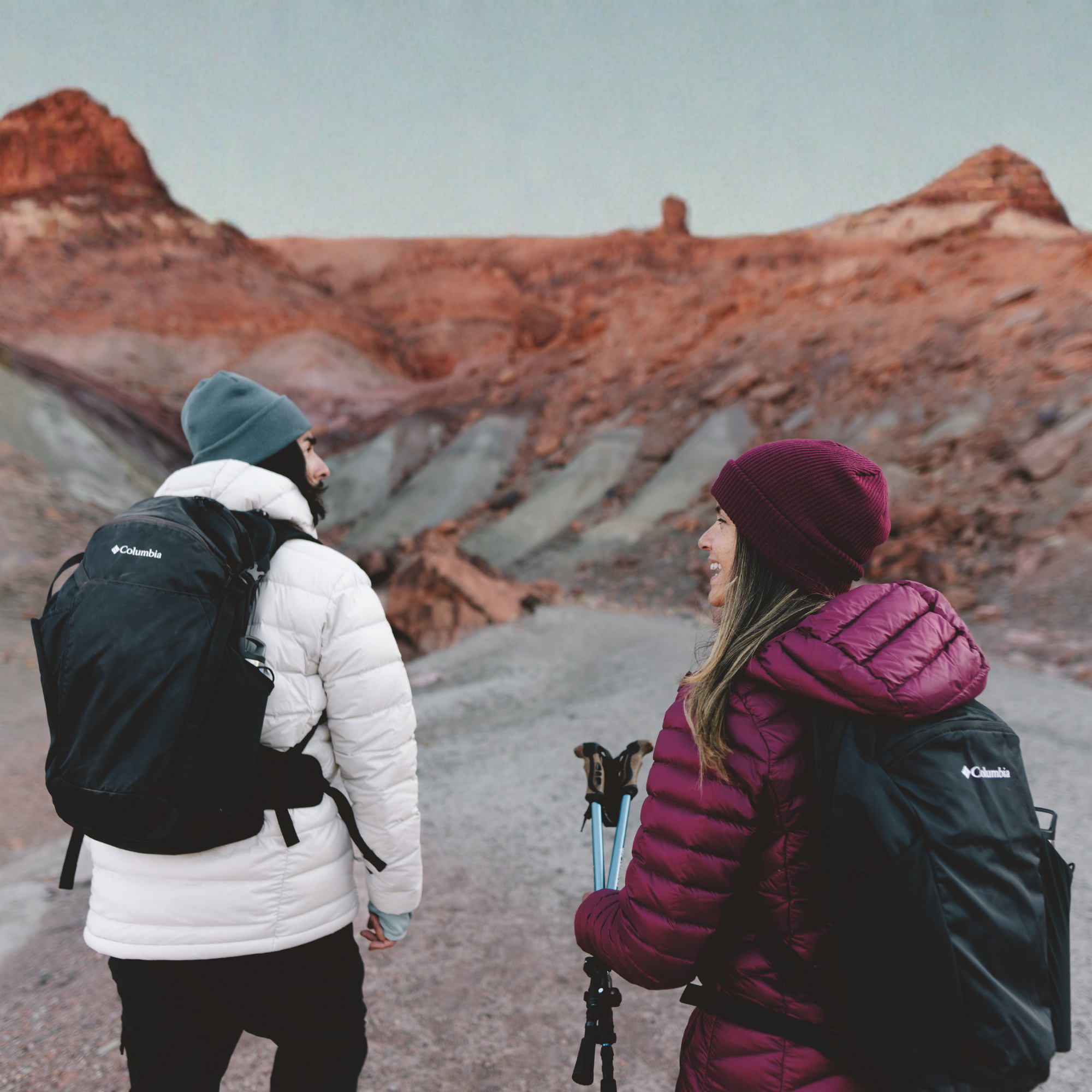 Two hikers wearing Columbia backpacks and winter clothing walk through a rocky desert landscape with reddish rock formations in the background using Columbia Blackcomb Ridge 18L Backpack Black 010 2121411