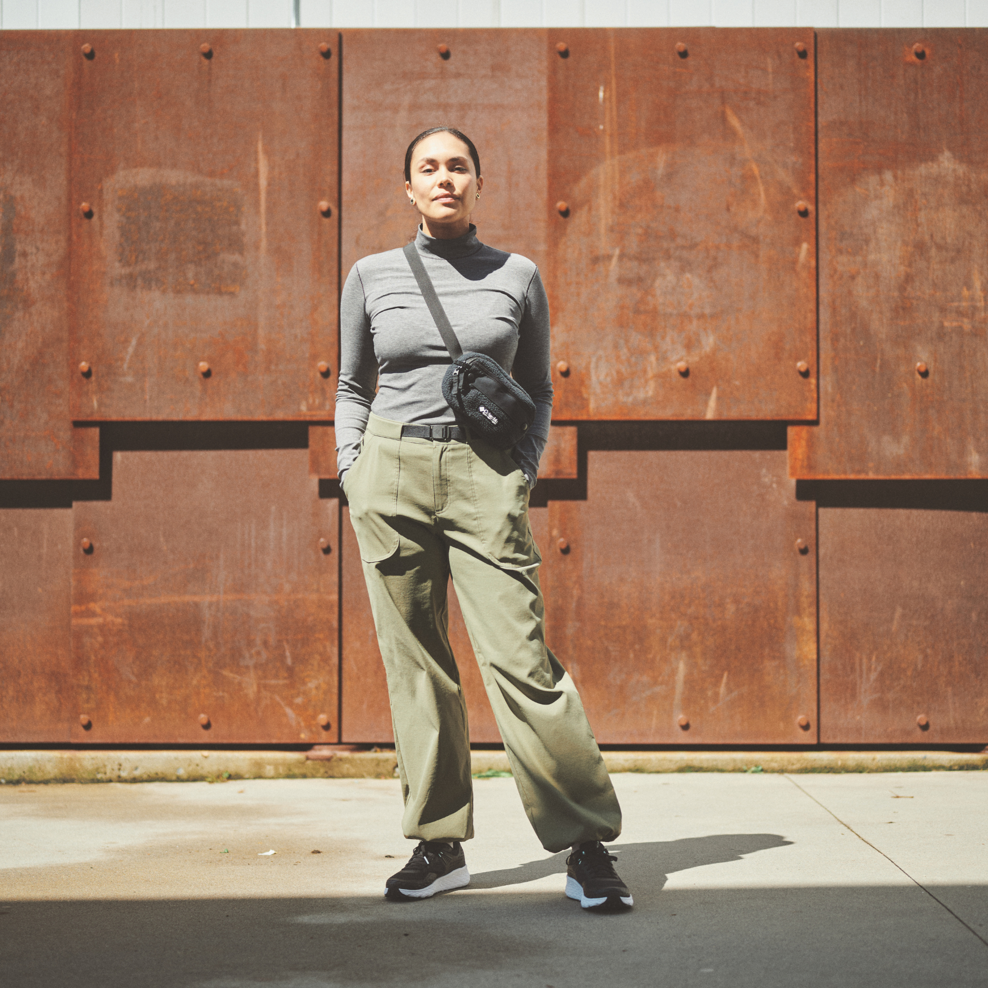 A woman stands outdoors in front of a large rusted metal wall, wearing a gray ribbed turtleneck top, olive green wide-leg cargo pants, black runners, and a Columbia Helvetia II Sherpa Hip Pack Black 010 2094541