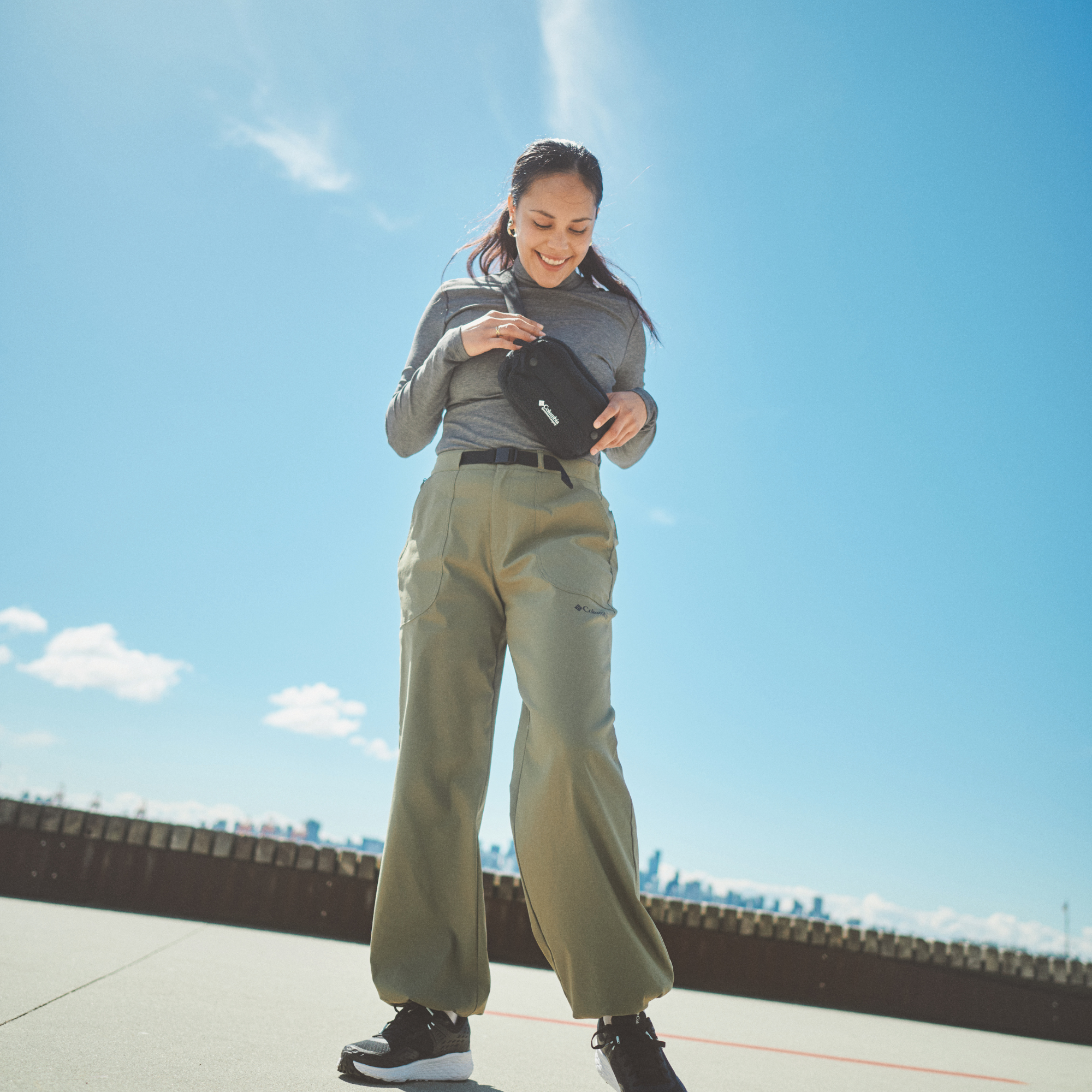 A woman outdoors under a bright blue sky wearing a grey long-sleeve top, olive green wide-leg cargo pants, and black sneakers, holding a Columbia Helvetia II Sherpa Hip Pack Black 010 2094541