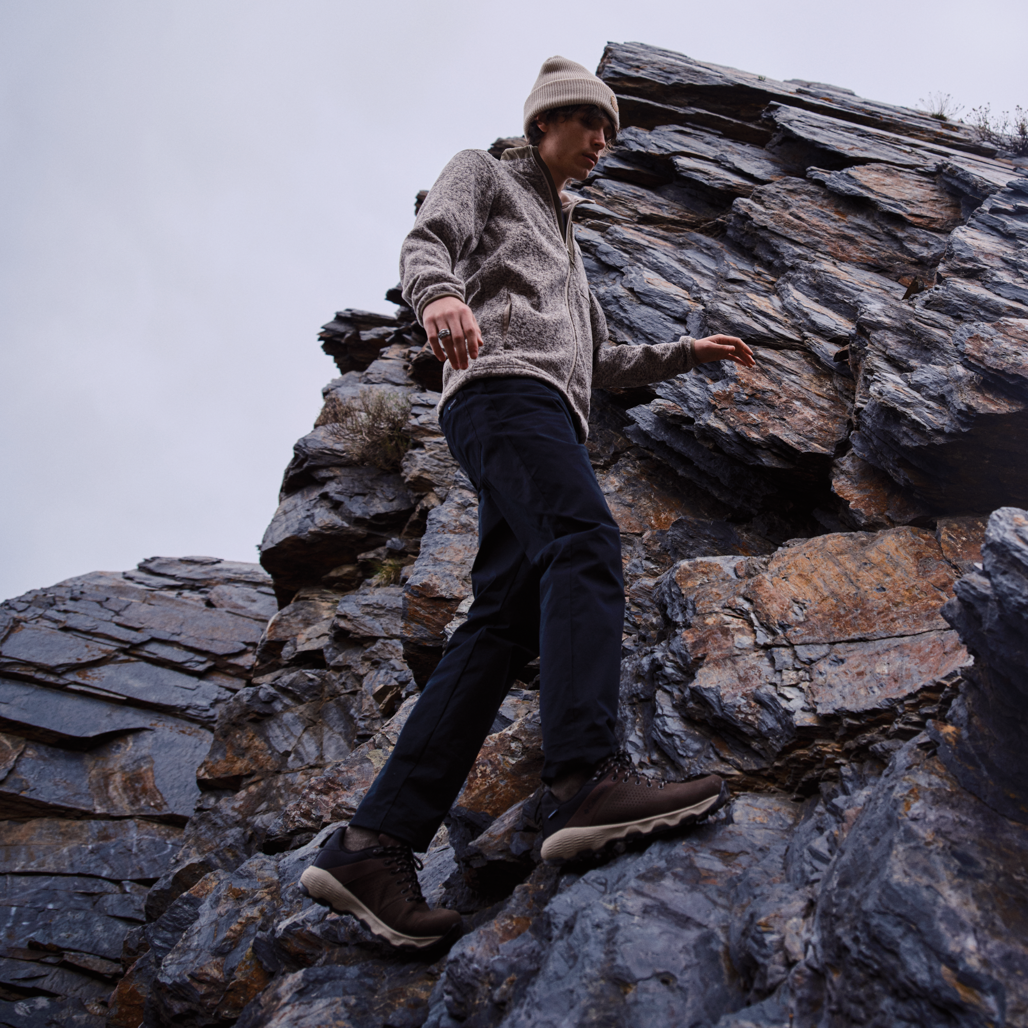 A man wearing a beige knit beanie, a textured gray zip-up hoodie, dark navy pants, and Columbia Men's Newton Nimble LTR Waterproof Trail Shoes Tundra Black 357 2140371 climbing a steep rocky cliff under an overcast sky.