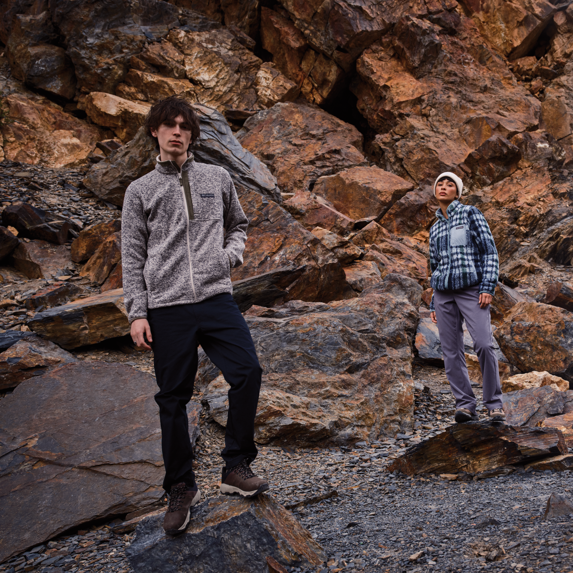 Two models standing on a rocky outdoor terrain, showcasing outdoor apparel. The model in the foreground wears a heather gray zip-up fleece, black pants, and Columbia Men's Newton Nimble LTR Waterproof Trail Shoes Tundra Black 357 2140371
