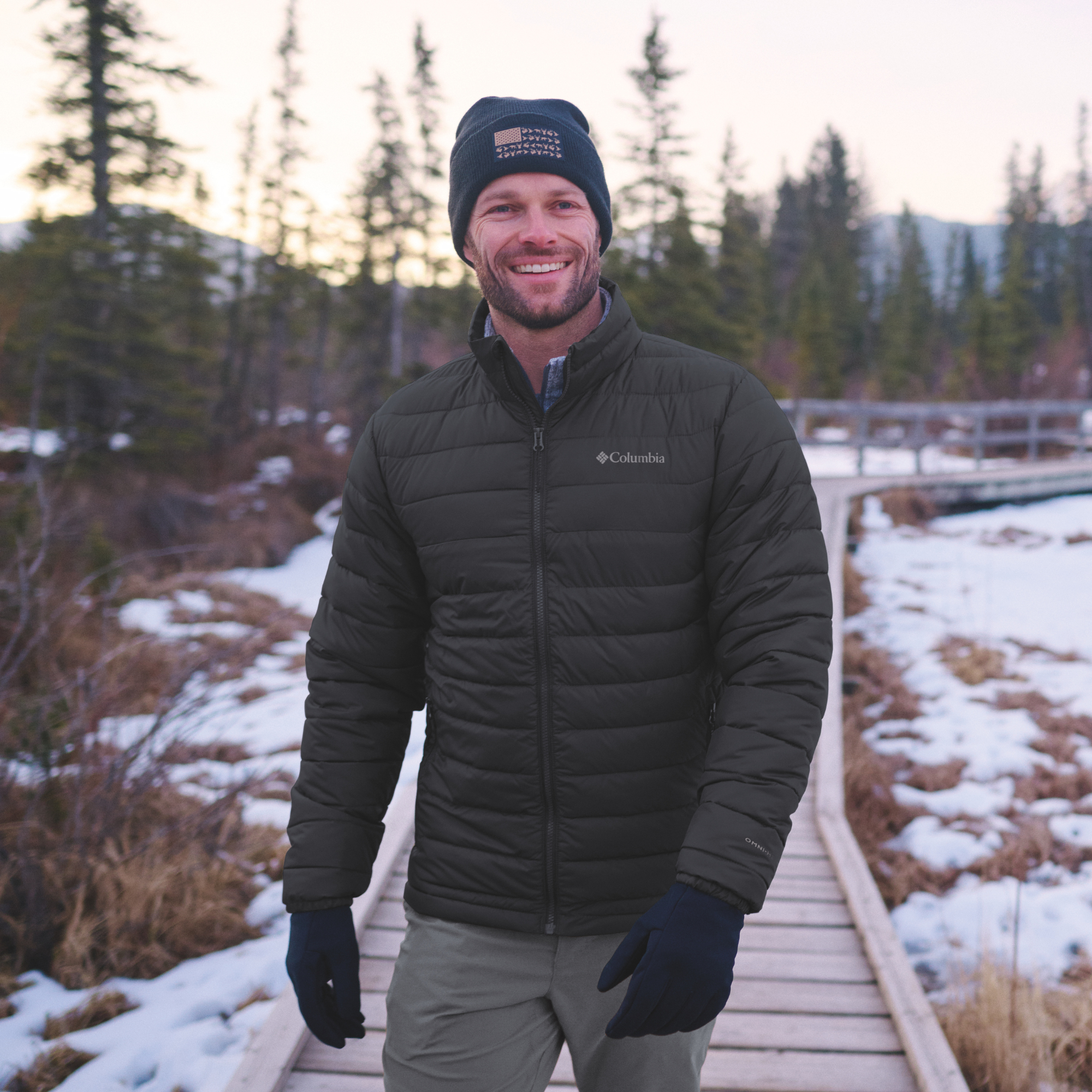 A man smiling outdoors on a wooden boardwalk wearing a Columbia Men's Powder Lite II Insulated Jacket Black 010 2086964, a black beanie, and black gloves, with snowy ground and evergreen trees in the background.