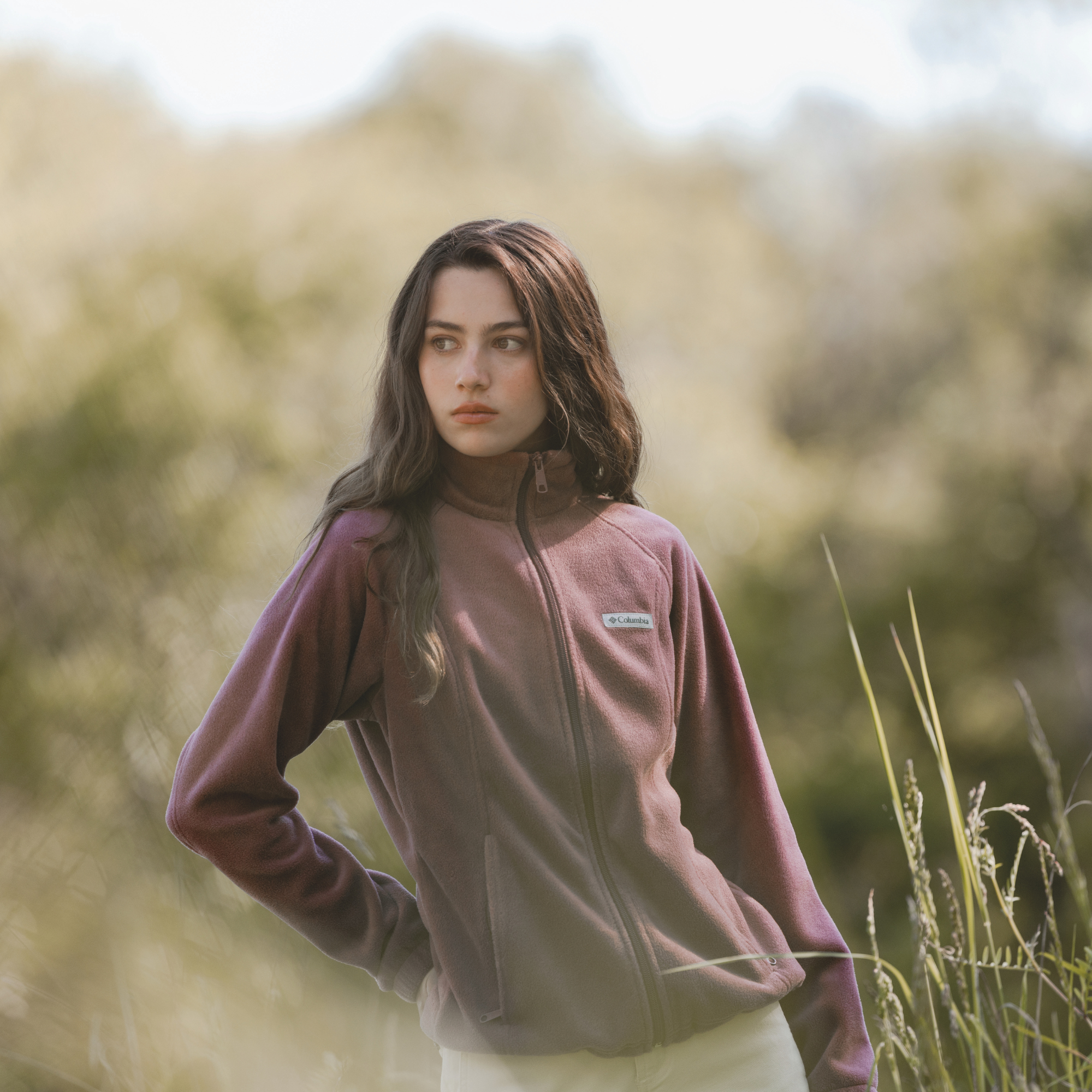 A woman wearing a Columbia Women's Benton Springs Full Zip Fleece Jacket Antique Mauve 553 1372111 stands outdoors in a grassy field with a soft-focus natural background.