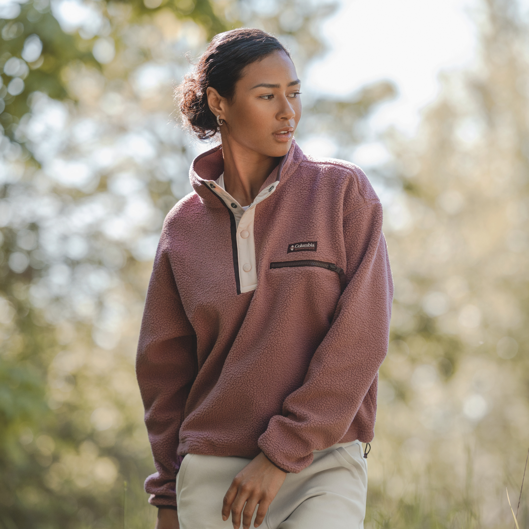A woman wearing a Columbia Women's Helvetia II Cropped Sherpa Half Snap Fleece Antique Mauve Mineral Pink 552 2092231 photographed outdoors against a soft, blurred natural background.
