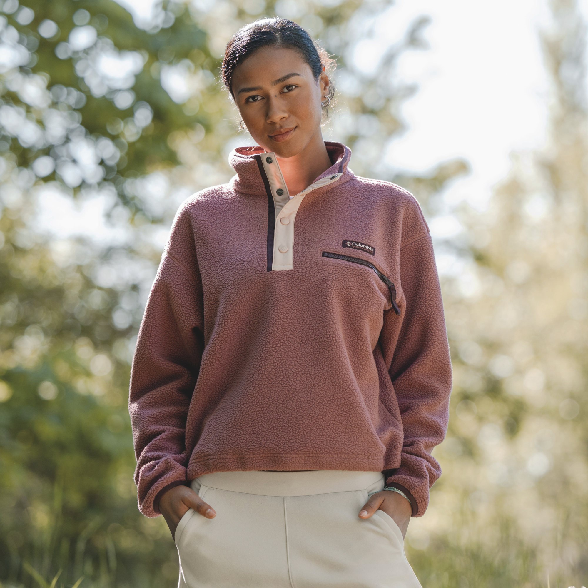 A woman wearing a Columbia Women's Helvetia II Cropped Sherpa Half Snap Fleece Antique Mauve Mineral Pink 552 2092231, standing outdoors in a sunny, blurred green environment.