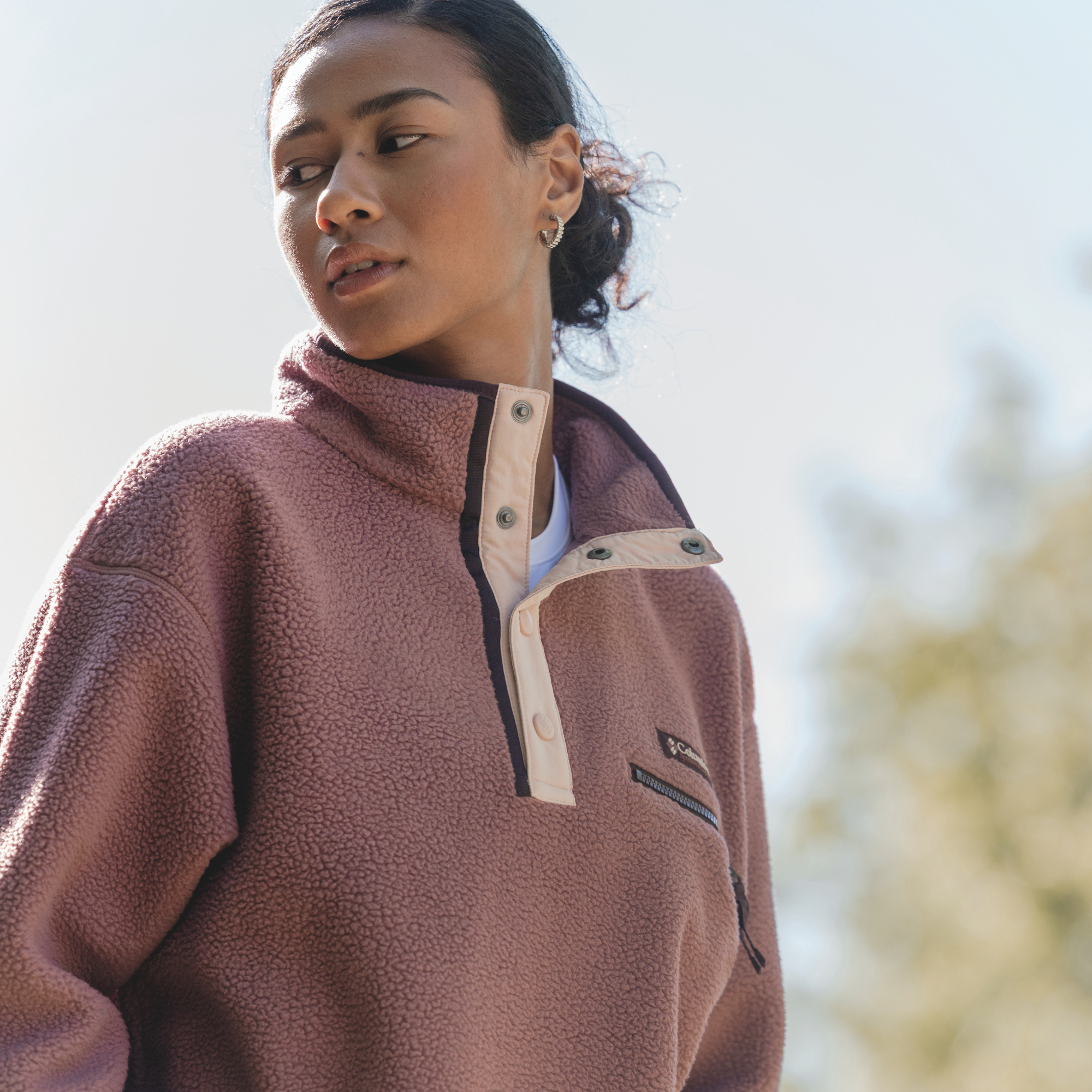 A woman wearing a Columbia Women's Helvetia II Cropped Sherpa Half Snap Fleece Antique Mauve Mineral Pink 552 2092231, photographed outdoors against a bright, blurred sky.