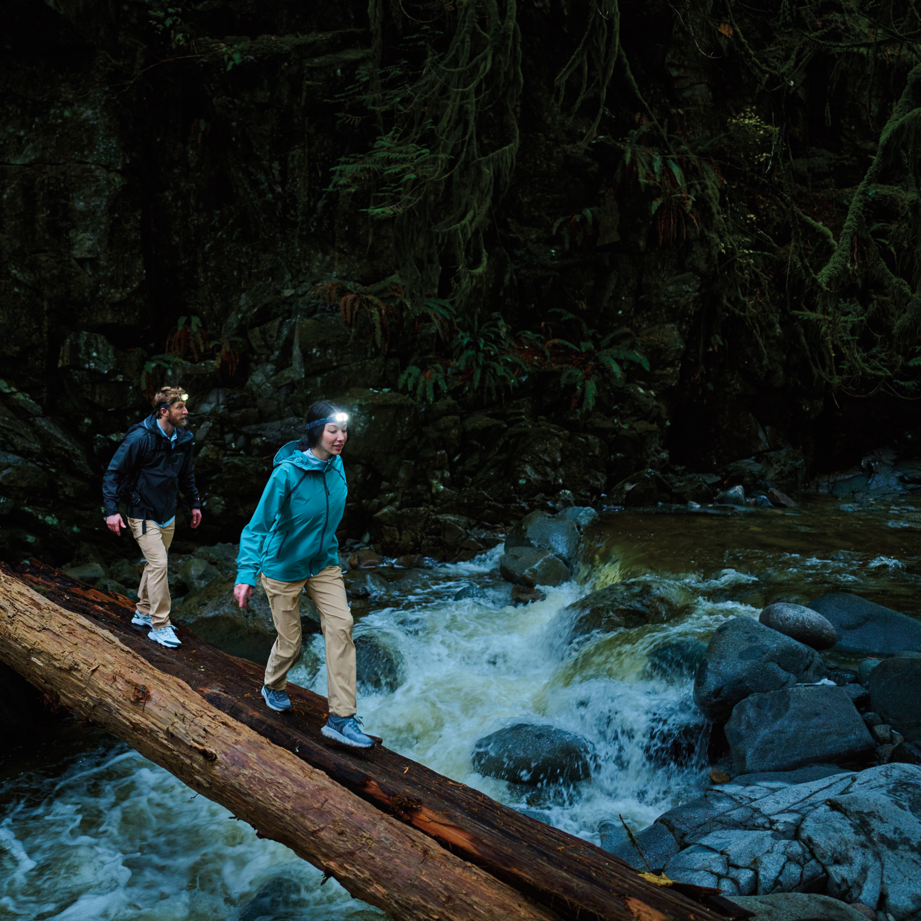 Two hikers hiking across a fallen tree over a river wearing waterproof jackets and headtorches. Woman wearing Columbia Women's Reign No Shine Outdry Extreme Waterproof Jacket River Blue 364 2116293.