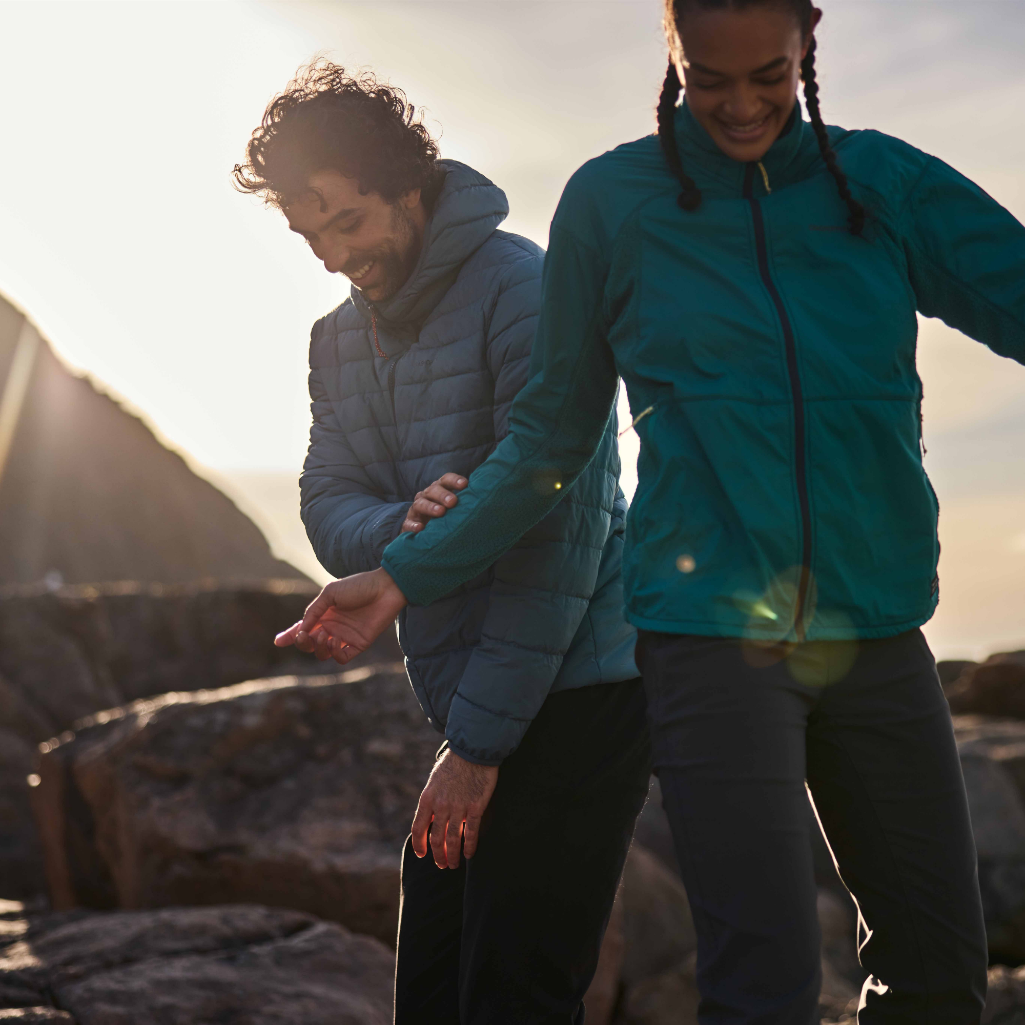 Two people walking outdoors on rocky ground at sunset, wearing lightweight jackets. One person wears a teal zip-front jacket, and the other wears a Craghoppers Men's Complite IX Hooded Insulated Jacket Winter Storm 1Y0 CMN323.