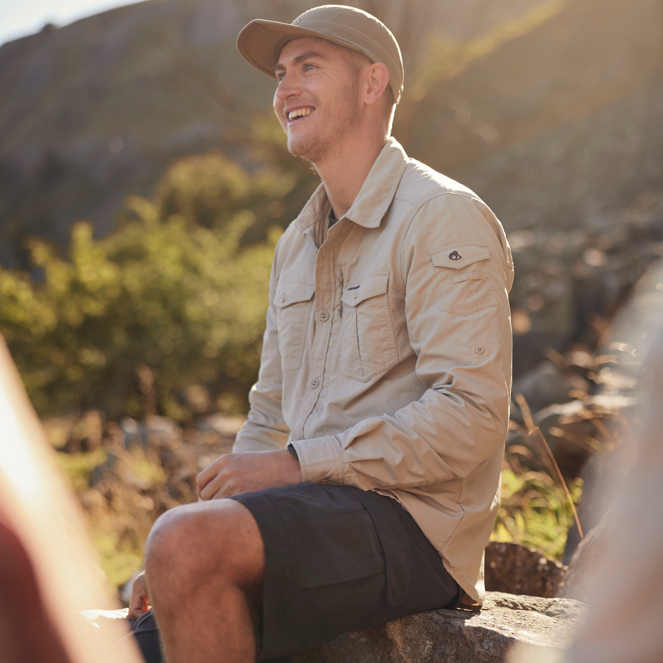 Man sitting outdoors in a natural setting, wearing a beige shirt, cap and Craghoppers Men's NosiLife III Cargo Shorts 10.5 inch Black Pepper 7J8 CMJ651