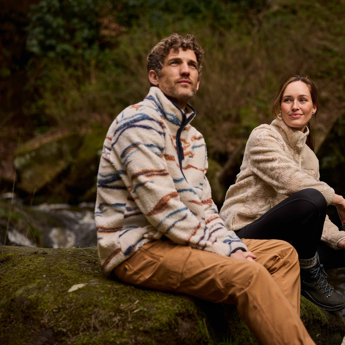 Man wearing Craghoppers Men's Rawthey National Trust Half Zip Fleece Top Ecru Winterstorm 1XJ CMA1425 sitting on a mossy boulder beside a stream with a woman in a sherpa fleece sitting beside him