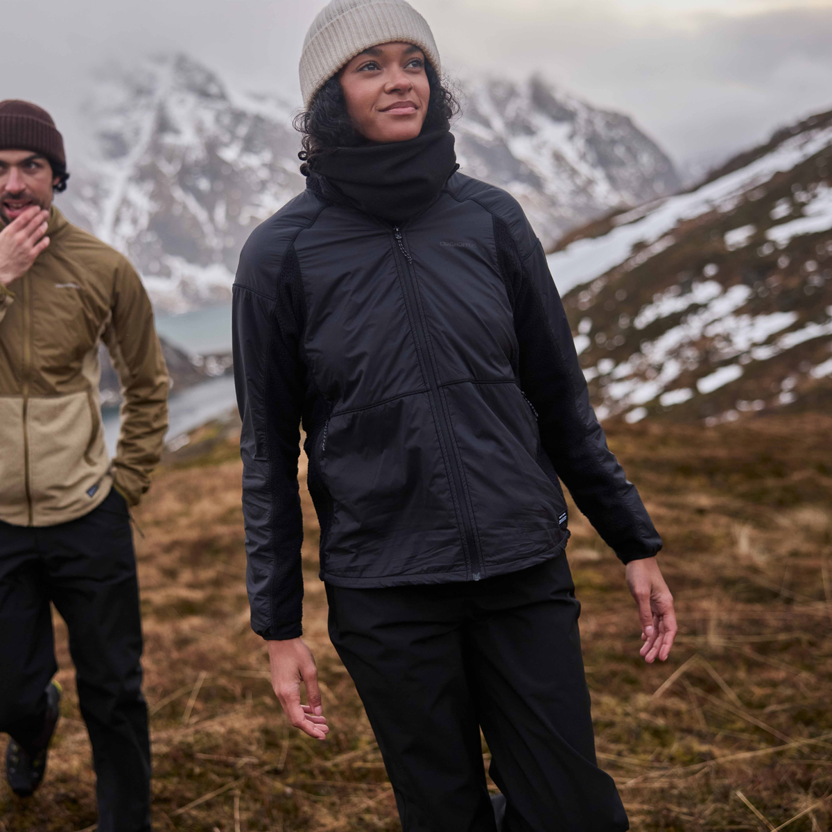 A woman wearing a black outdoor jacket and Craghoppers Women's Aysgarth Pro Thermo Waterproof Trousers Black 800 CWW1228 stands in a mountain landscape with snow patches, while a man in a tan jacket walks in the background.