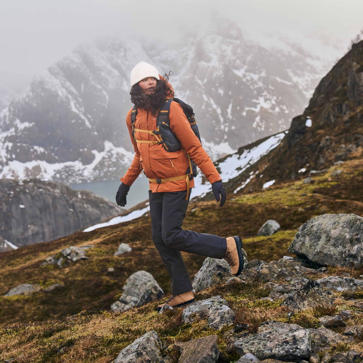 A person hiking on a rocky mountain slope wearing an orange puffer jacket, Craghoppers Women's Aysgarth Pro Thermo Waterproof Trousers Black 800 CWW1228, a white beanie, gloves, and a backpack, with snowy mountains in the background.