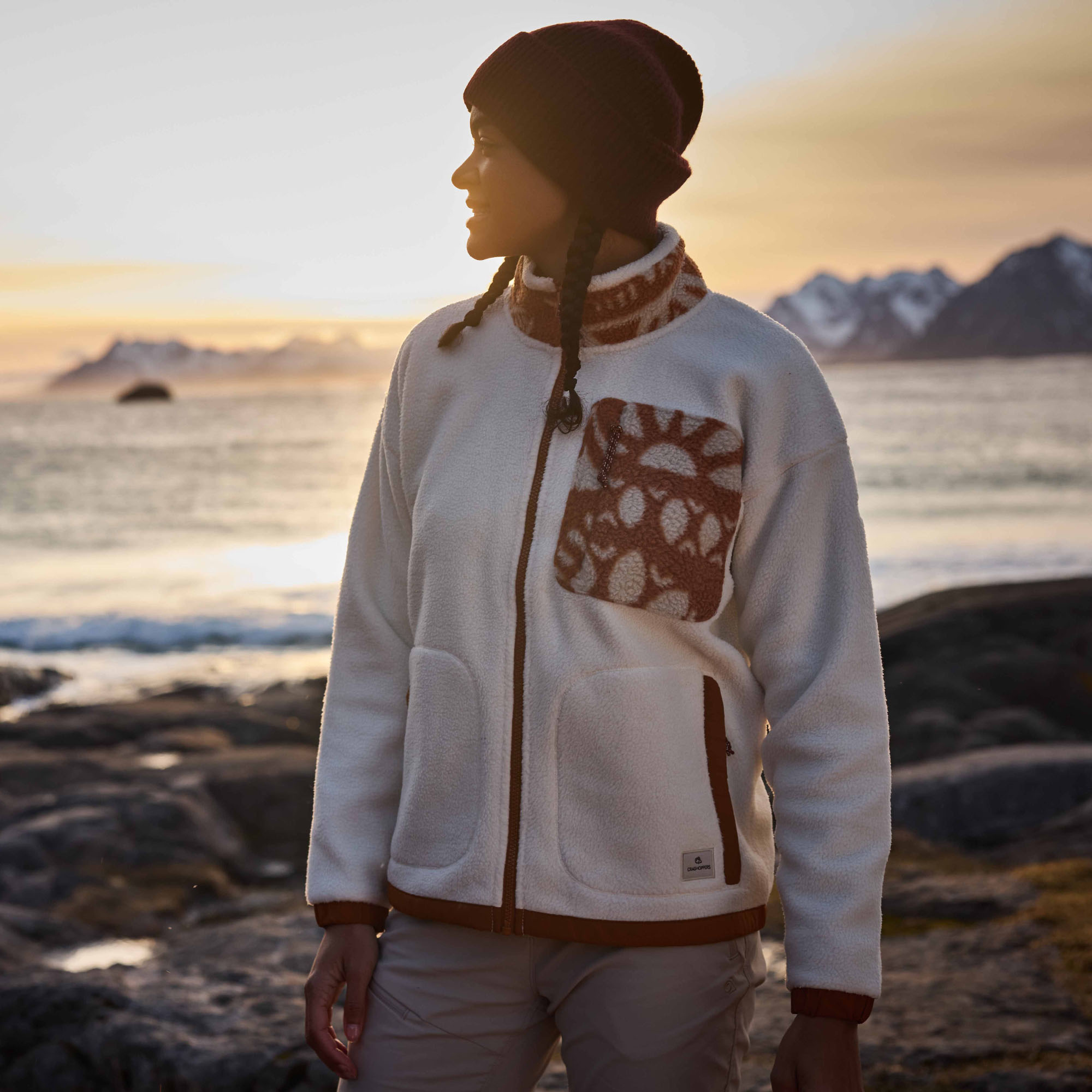 A woman wearing a Craghoppers Women's Sanaa Full Zip Fleece Ecru Hazel 2QF CWA436 stands on a rocky shoreline at sunset, with water and mountains in the background.