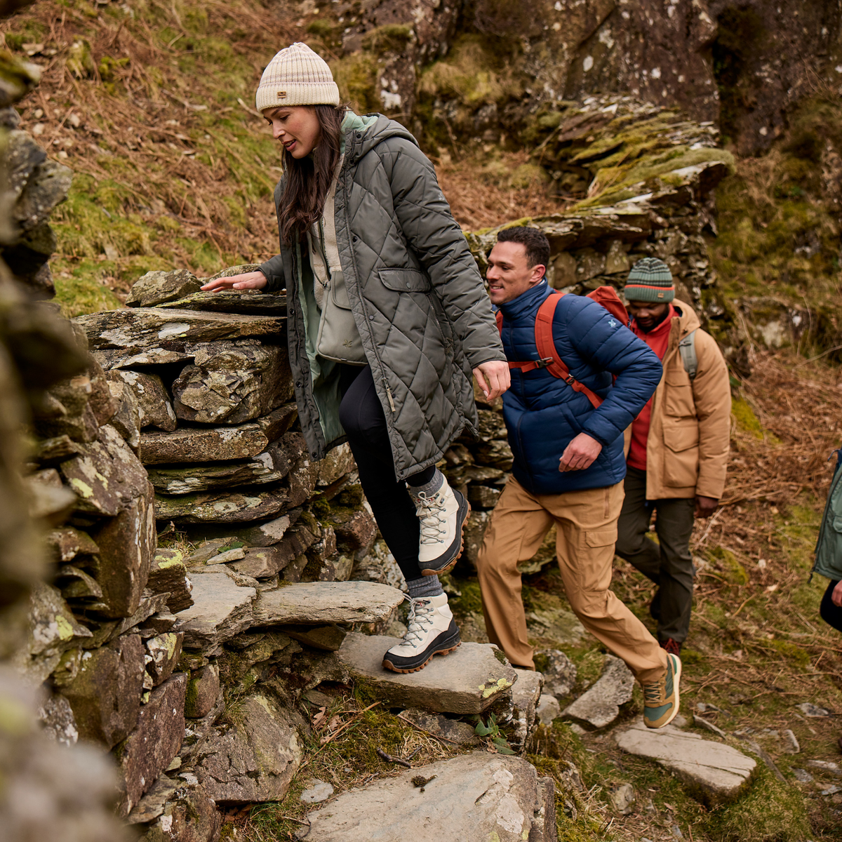 Three people hiking up stone steps in a rugged outdoor mountain setting, wearing insulated jackets and outdoor clothing. Main woman in image wearing Craghoppers Women’s Sweetgrass National Trust Insulated Parka Nori Green 1KZ CWN309.