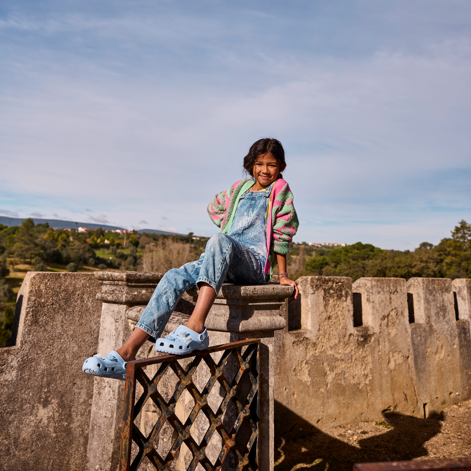 Young girl sititing on a pillar wearing Crocs Classic Clogs Junior Blue Calcite 4NS 206991