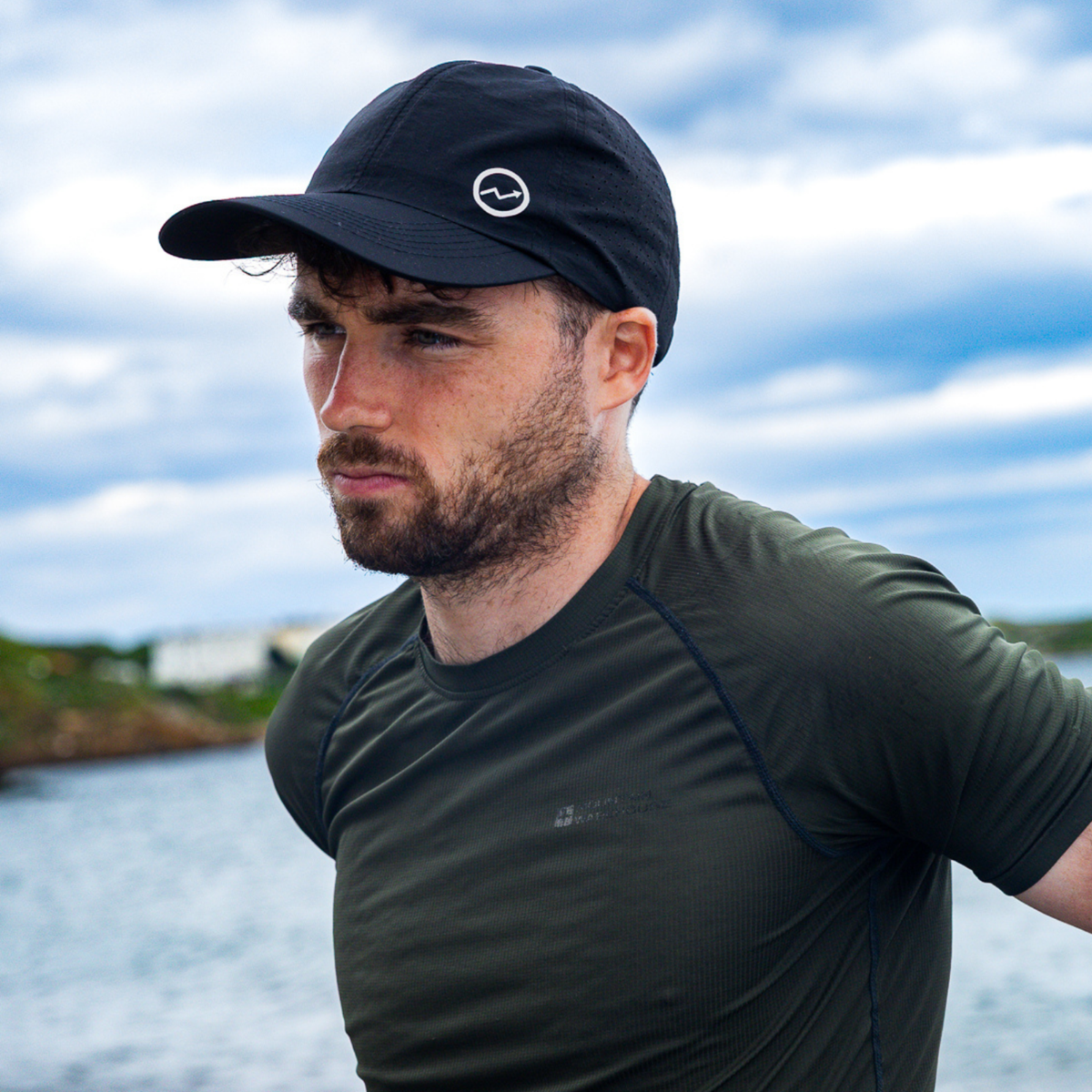 A man wearing a Eos Elite Unisex HighER Running Cap Black EOS-CAP1 and an olive green athletic shirt, photographed outdoors with water and a cloudy sky in the background.