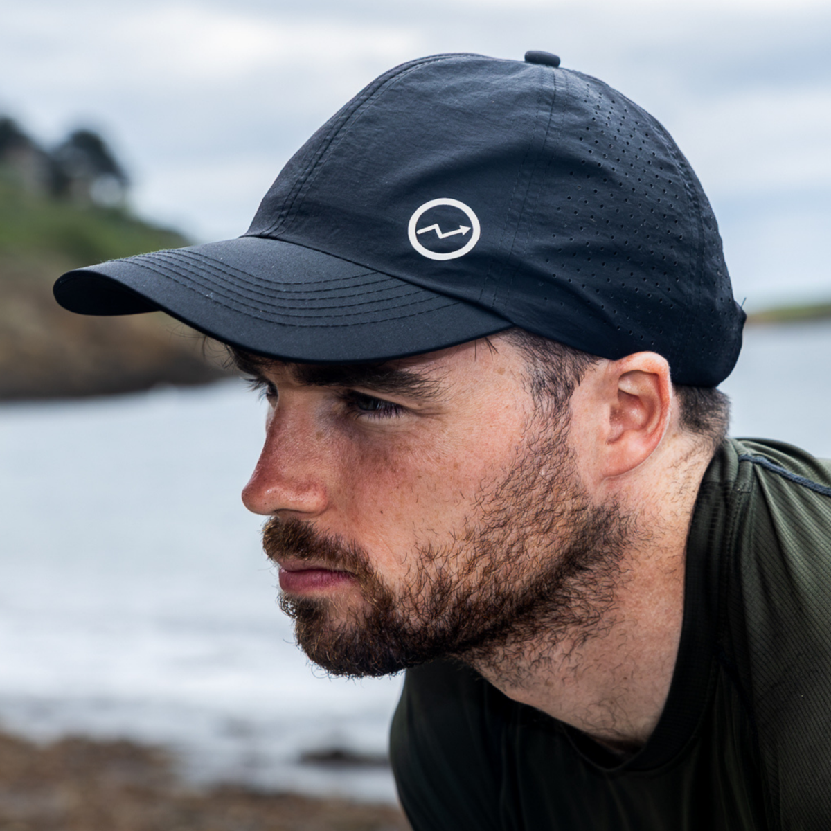 A man in profile wearing a Eos Elite Unisex HighER Running Cap Black EOS-CAP1, photographed outdoors near water and rocks.
