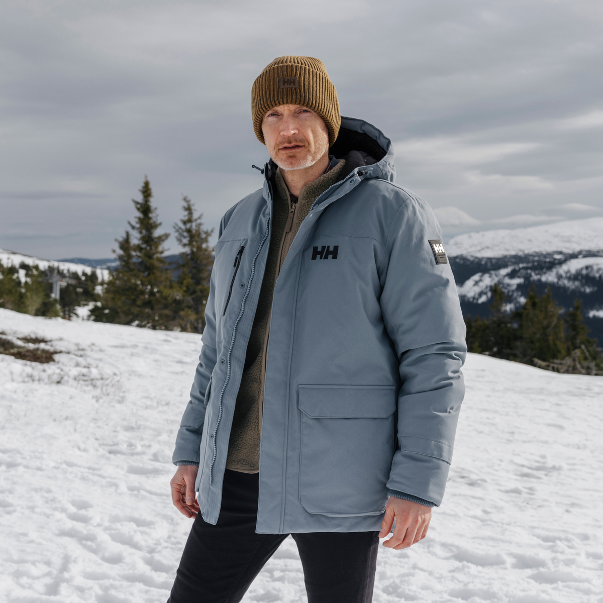 Man wearing a blue jacket with a Helly Hansen Unisex HH Rib Beanie Hat Sepia 718 54020 in a snowy landscape